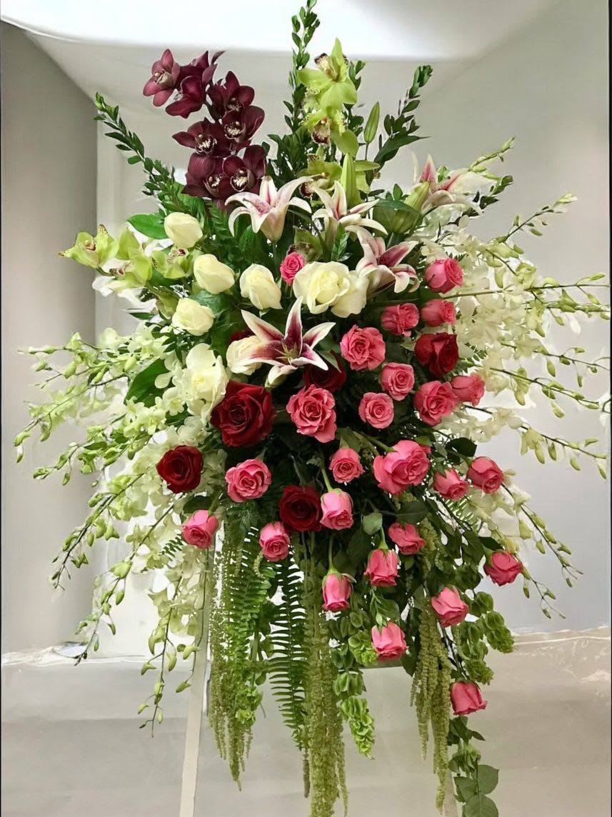 Tall standing floral spray with red and pink roses, lilies, orchids and trailing greenery.