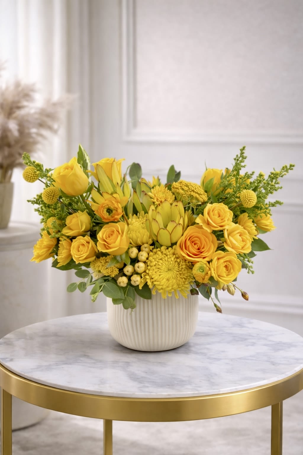 Low yellow flower arrangement with roses, protea, and mums in a white ceramic pot on a marble table