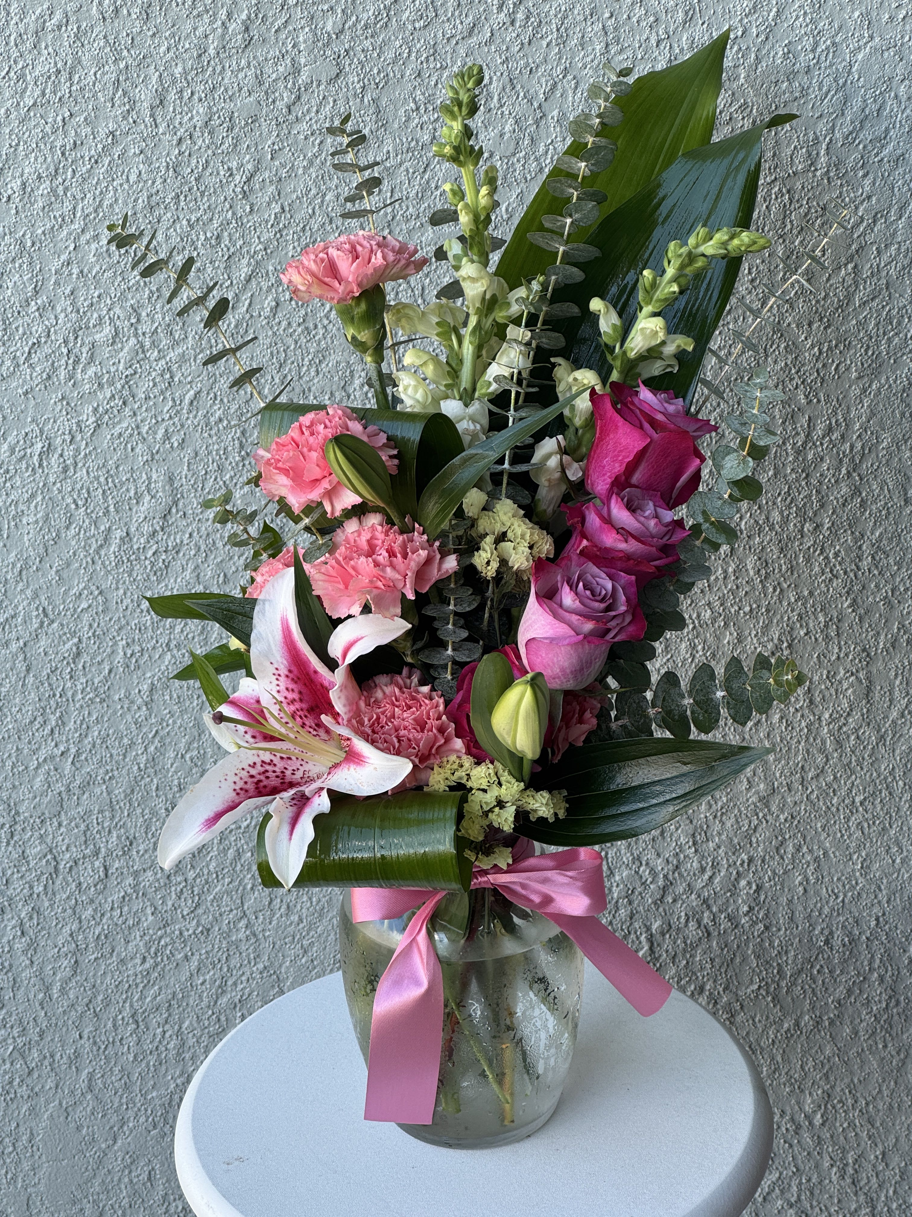 Tall pink and white arrangement with roses, lilies, and carnations in a glass vase with a pink ribbon.