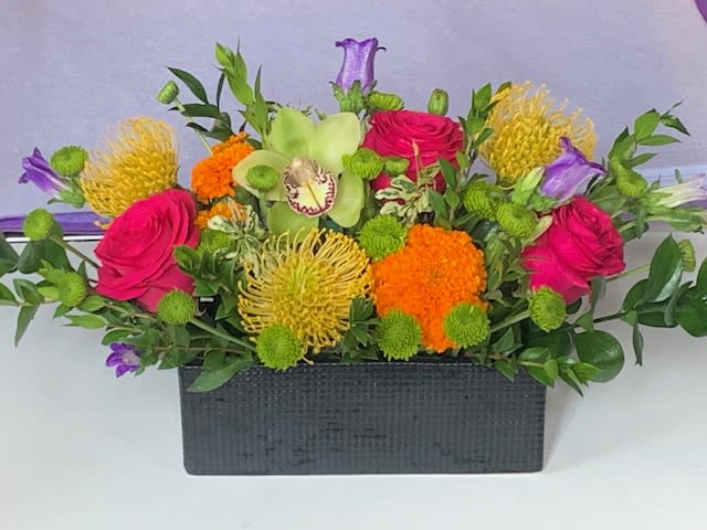 The Southern Tropical - Long and low vase featuring an electric mix of bright summery tropical vibes. Flowers featured include pin cushion protea, orchids, marigolds and hot pink roses.