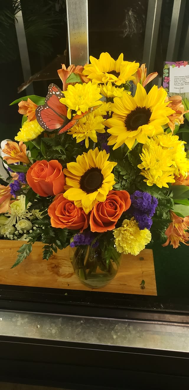 Hello Ms Lena - Golden sunflowers steal the show in this mixed-color flower arrangement, a cheerful birthday bouquet or Just Because gift that brings sunny warmth to Mother's Day, get well wishes, congratulations, or a heartfelt thinking-of-you moment. The upright clear glass vase and playful orange butterfly pick give the design a garden-fresh, celebratory feel that also suits housewarming, graduation, anniversary, or seasonal Easter displays.  Clusters of large yellow sunflowers are paired with bright orange roses, orange alstroemeria, peach carnations, yellow cushion-style blooms, and purple filler flowers for striking contrast and texture. The clear glass vase reveals the stems for a classic silhouette and easy placement on a table or mantel; the size reads airy and upright, ideal for sending sympathy or funeral condolences when a bright, comforting touch is wanted as well as for festive holidays like Thanksgiving, Christmas, or Fourth of July. Simple care-fresh water and a cool spot-will keep the bouquet looking vibrant for days.