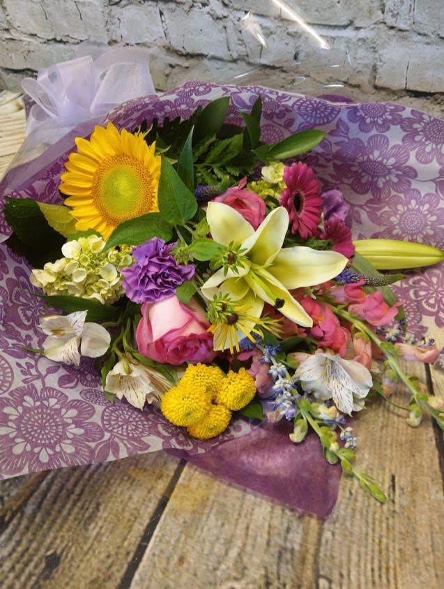 Mixed bouquet with a sunflower, lilies, roses, and purple blooms wrapped in purple paper