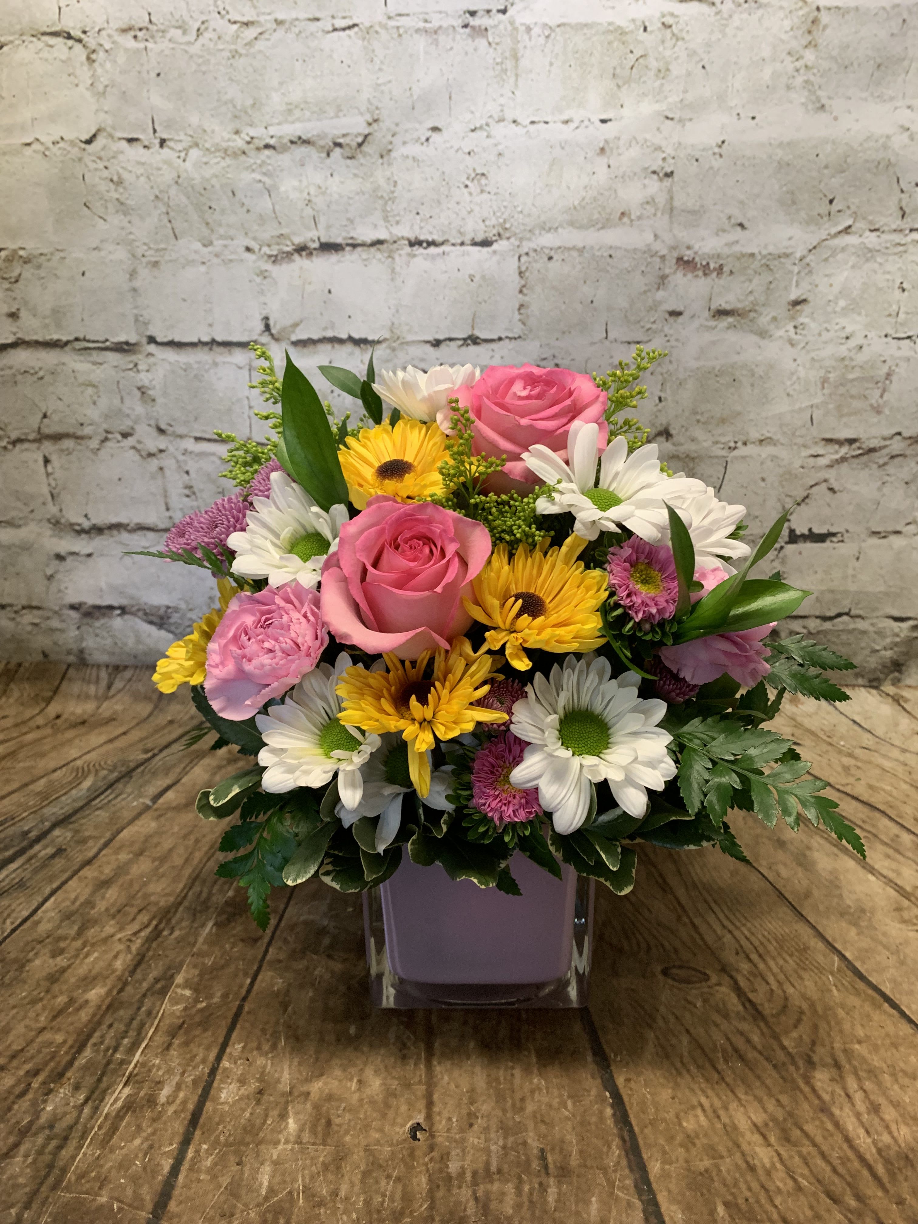 Mixed bouquet of pink roses, daisies, and yellow flowers in a square vase