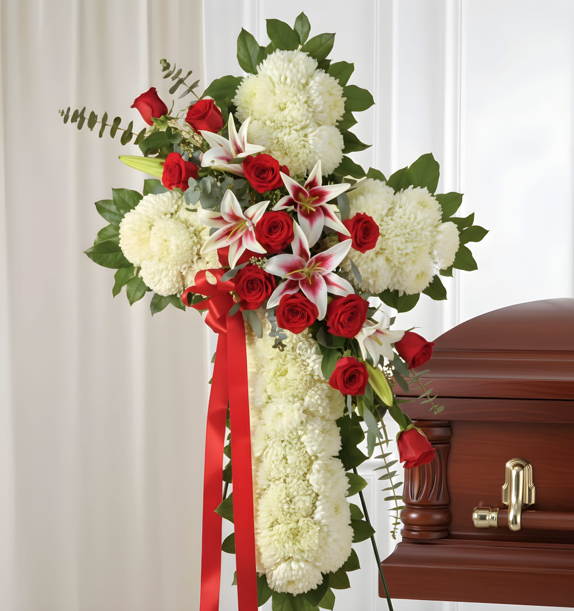 Elegant Standing Cross (Roses &amp; Lilies) - Dense white carnations shaped into a dignified standing cross create a solemn, reverent tribute perfect for a funeral or memorial service. This funeral spray combines that pure white base with clusters of red roses and white lilies - the red ribbon and lush green foliage heighten the contrast so the person you're honoring will feel remembered with both grace and heartfelt color.  Designed for upright display on a standing easel beside a casket, the arrangement uses a floral cross frame to achieve a large-scale, traditional presentation. The composition features tightly grouped carnations for a clean silhouette, red roses and lilies arranged across the top and center for visual focus, and a long red ribbon as a respectful accent. Offered as a standing spray suitable for services, it arrives ready to place on the easel for an immediate, dignified statement.