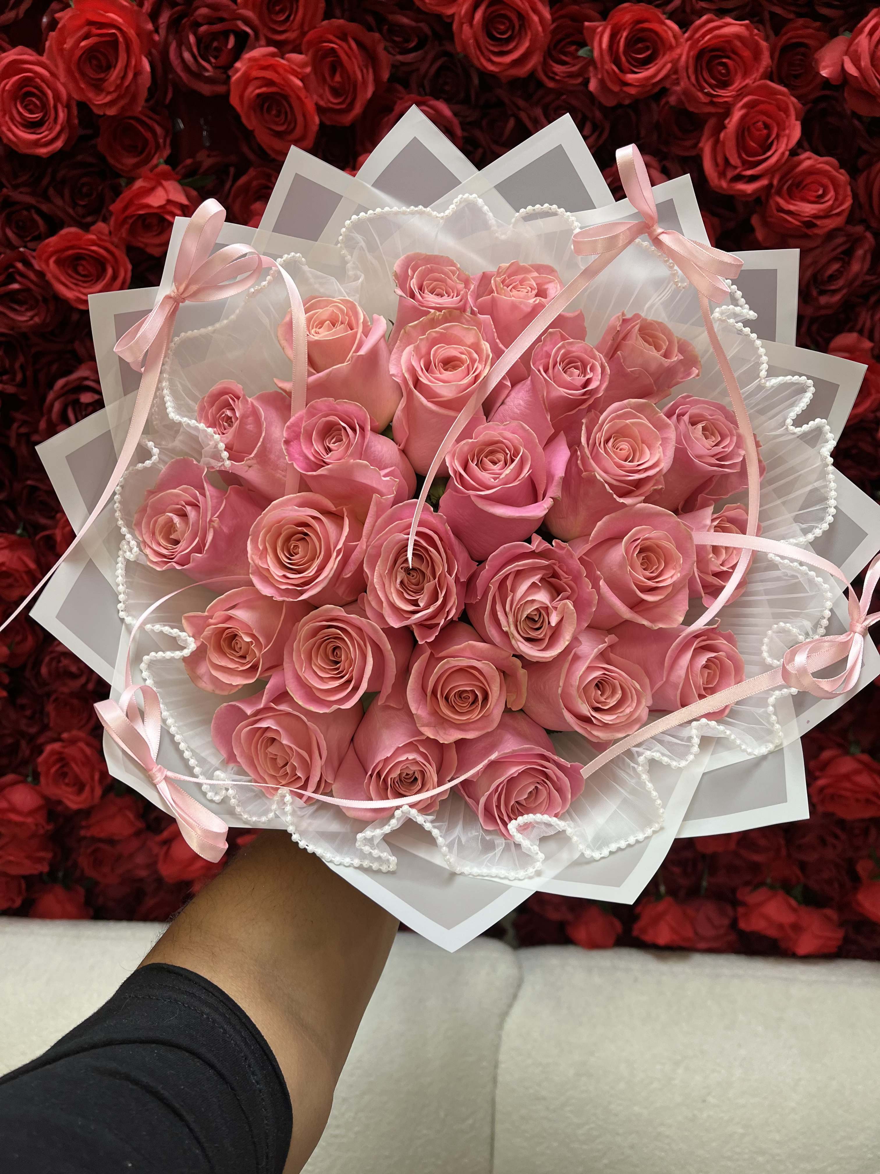 Handheld bouquet of pink roses wrapped in white paper with ribbons and pearl trim