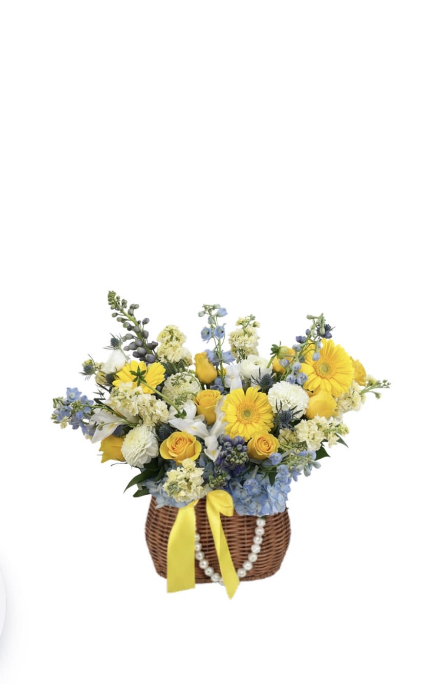 Dolce Vita Basket - A cheerful, garden-inspired basket arrangement featuring yellow gerbera daisies, roses, stock, blue delphinium, hydrangea, white ranunculus, and carnations. Designed with a soft blend of warm and cool tones, this piece brings a fresh, elegant feel that’s perfect for any occasion.