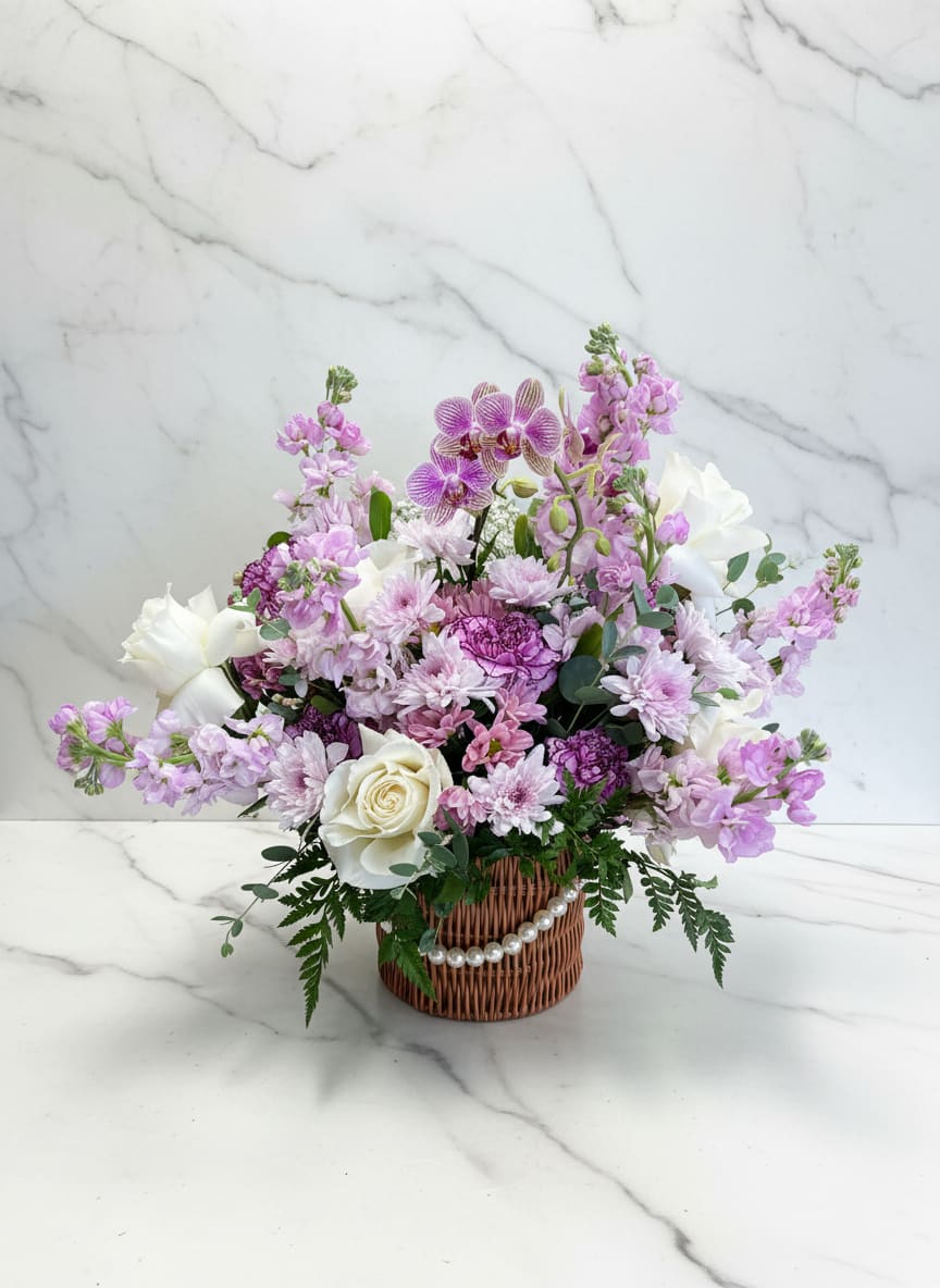 Lavender Orchid Basket - A soft and elegant round wicker basket featuring a mini double orchid plant as the focal point. Designed with white roses, a bicolor purple carnation, lavender stock, lavender daisies, mums, and fresh greenery, this arrangement blends gentle lavender tones with crisp whites for a balanced, garden-inspired look. Perfect for a calming, refined gift. 