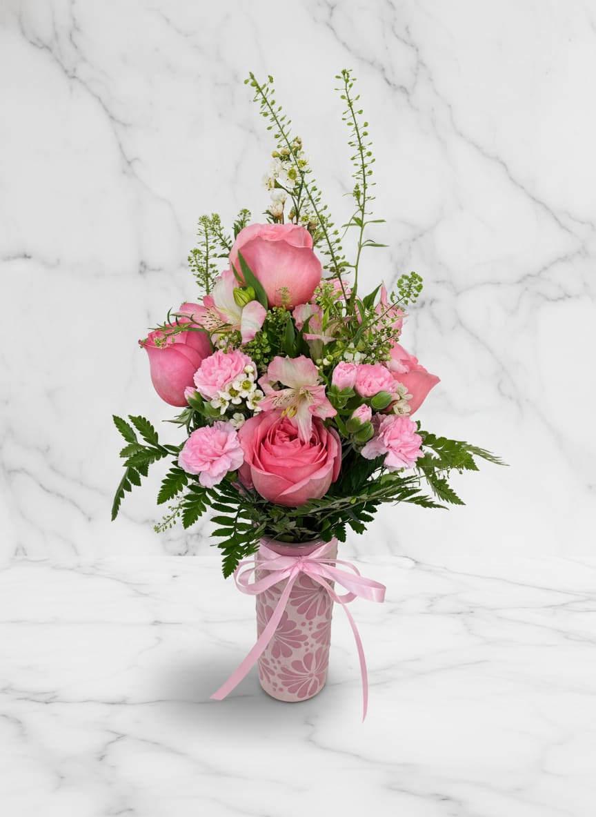 Cielito lindo Cup No.2 - Arranged in a patterned pink ceramic vase and finished with a satin pink ribbon, the design is gift-ready and easy to display. The mix includes roses, carnations, alstroemeria touches and small white filler blooms for contrast, with greenery that adds texture and movement. The compact base rising to a softer, taller top gives it presence on a bedside table, mantel, or office desk without overwhelming the space. 
