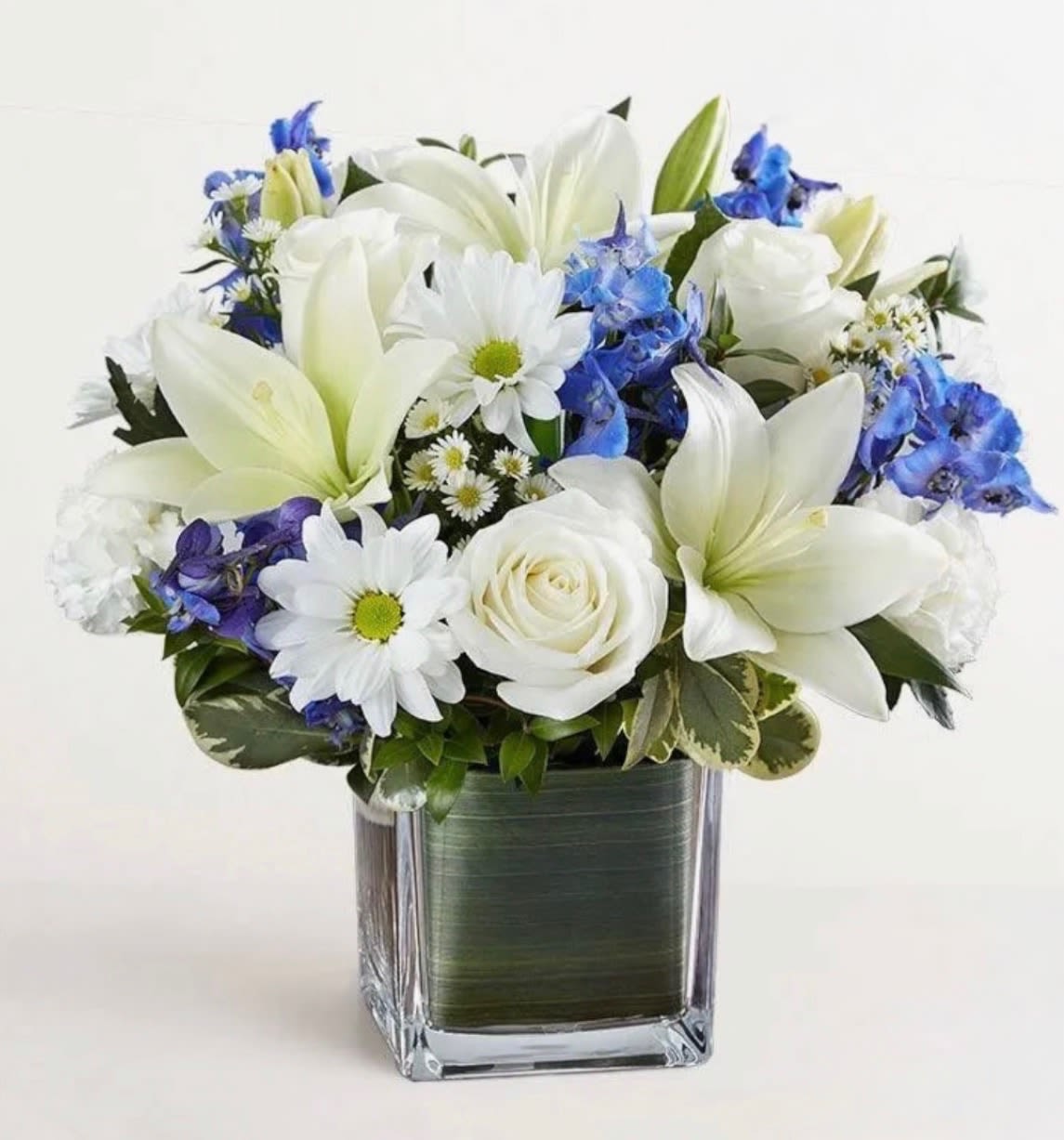White lilies, roses, and daisies with blue flowers in a square glass vase