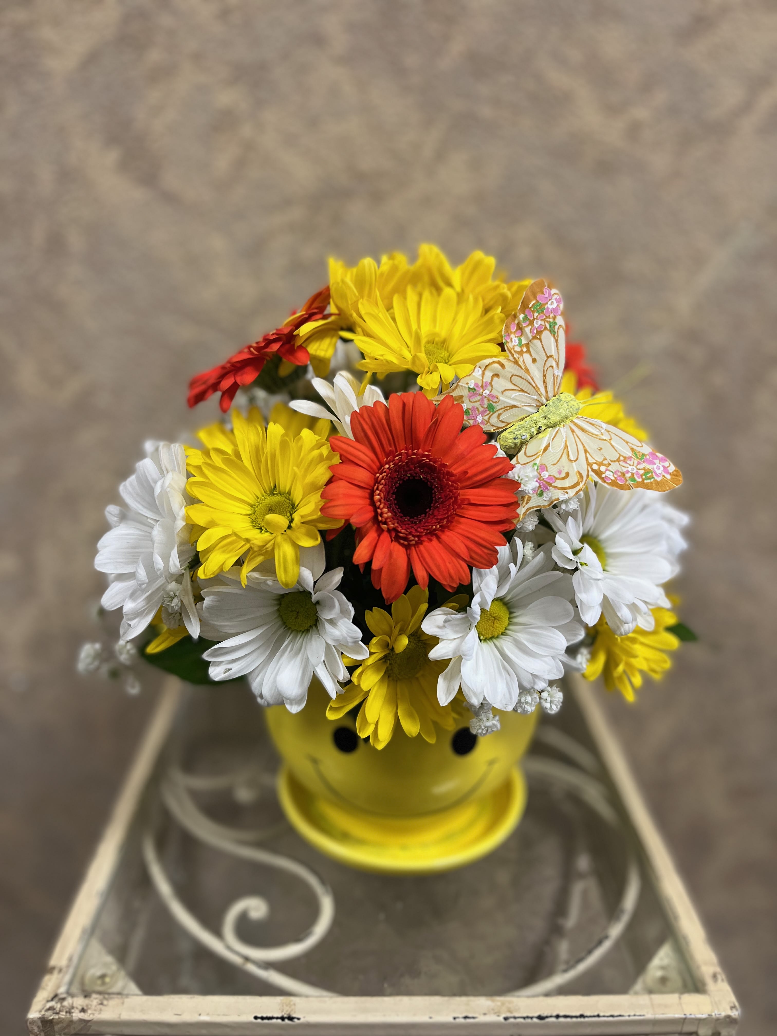 Colorful daisy bouquet in a yellow smiley-face pot with a decorative butterfly