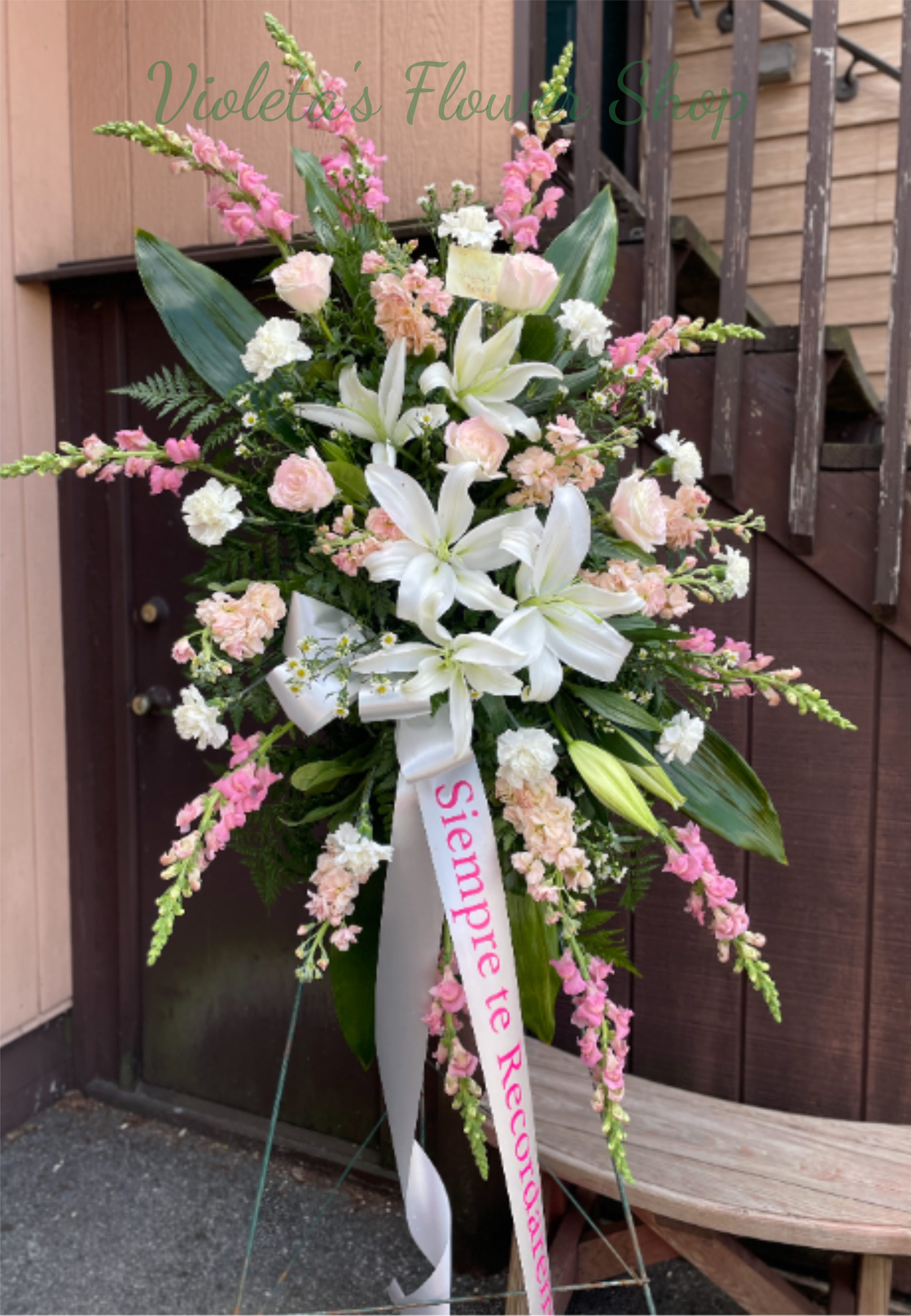 Treasure Memories Standing Spray - Large white lilies take center stage in this sympathy standing spray, creating a soft, elegant tribute for a funeral or memorial service. Designed as a vertical flower arrangement for remembrance, the pale pink roses and carnations surround the lilies while tall pink snapdragon spikes add graceful height-together the mix reads as gentle, consoling, and reverent for the person you're honoring.  This arrangement uses white, pink, green and mixed tones against broad dark green foliage that gives a lush, full backdrop to the lighter blooms. Daisies and additional filler blossoms add texture and softness among the main lilies, roses, carnations, and snapdragons. The spray is finished on a standing easel with a white ribbon and a printed memorial sash for a formal, respectful presentation appropriate at a service.  For someone who needs a meaningful expression of sympathy, this standing spray says "remembered" with quiet beauty-the kind of floral tribute that comforts family and friends. Available ready to place on a funeral easel; please tell us the memorial wording for the printed sash when you order so we can personalize it for the service.