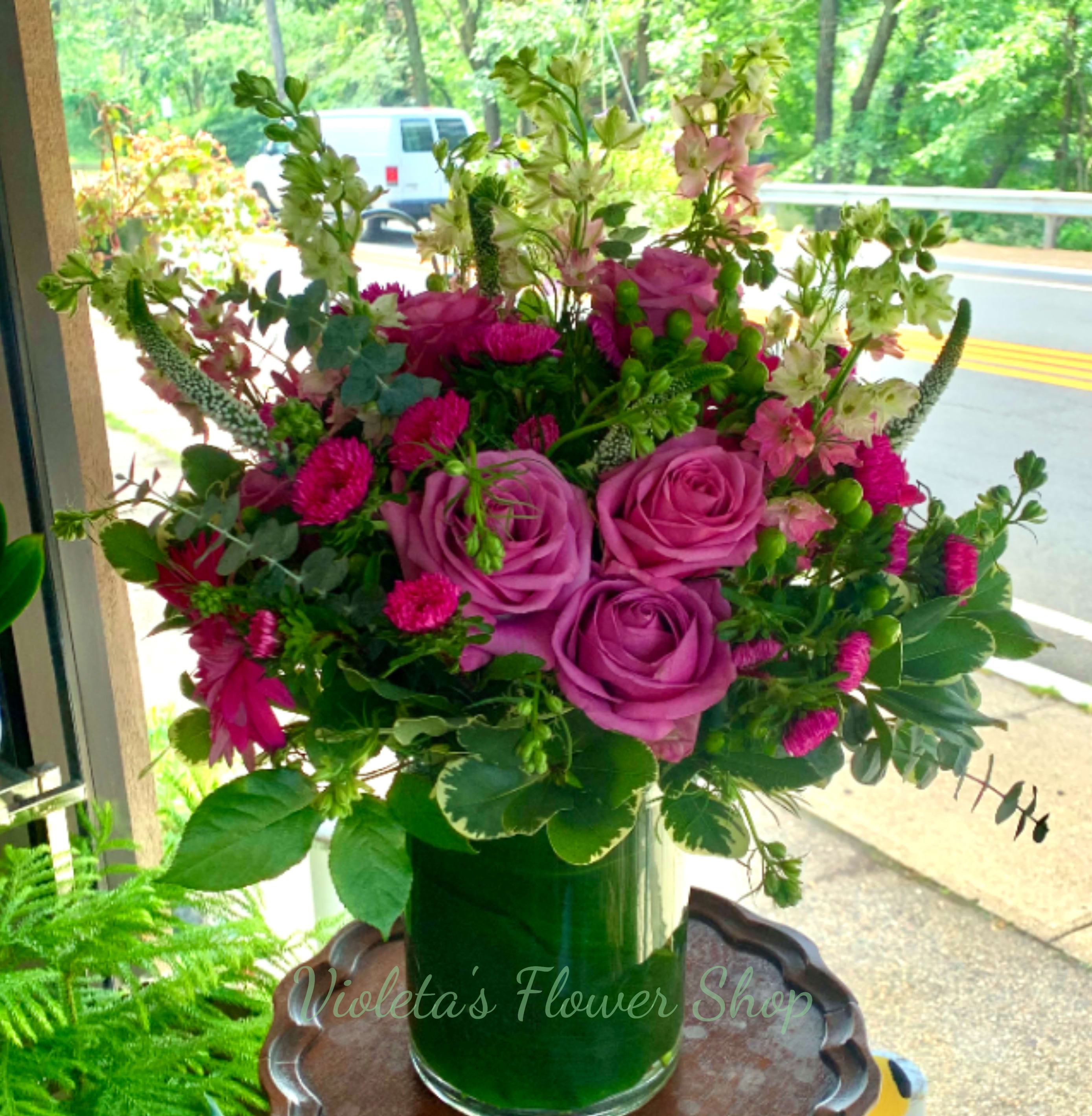 Raspberry Moody Arrangement - Lavender roses take center stage in this Raspberry Moody arrangement, their soft purple glow balanced by bright pink gerberas and tall pink larkspur spires for a lively, garden-style birthday bouquet or just-because surprise. Designed for housewarming, summer celebrations, or a heartfelt thank-you, the mix of rounded rose heads, textured asters, and airy stems reads romantic yet fresh-the kind of arrangement that makes the person you're thinking of feel truly celebrated.  Arranged in a clear cylindrical glass vase that reveals the lush green stems and water, this full, rounded composition brings a polished, upright presence to any room. Colors include pink, purple, white, and green with roses, asters, larkspur, daisies and complementary filler flowers creating layered texture and movement. It's an excellent pick for Mother's Day or Women's Day and for sending warm thinking-of-you or thank-you wishes; for best reception place in fresh water away from direct heat and enjoy its cheerful impact all season long.