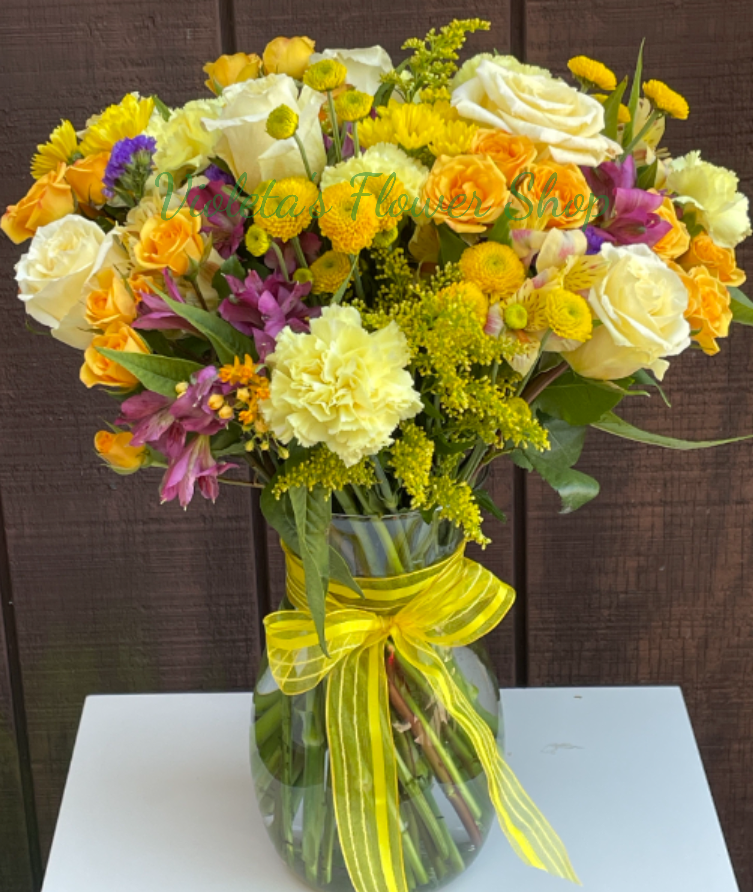 Field of Sunshine Arrangement - Sunny, layered color is what sets this Field of Sunshine arrangement apart - a bright, full bouquet perfect as a summer gift or a cheerful get-well surprise. The combination of yellow and white roses with yellow spray roses and pale carnations creates an immediate warm, uplifting impression, while purple alstroemeria threads in a flattering contrast that makes the colors pop. Tied with a bright yellow ribbon, it reads joyful and hopeful - the kind of arrangement that makes the person you're thinking of feel genuinely seen and cheered.  This design sits in a clear glass vase and features yellow button chrysanthemums and small yellow filler blooms for texture and a slightly rustic, garden-fresh feel. Dimensions are a full, rounded bouquet that works well on tabletops or bedside tables; stems are arranged for a balanced, layered look so blooms show from every angle. Ideal for summer birthdays, thank-you gestures, Día de las Flores Amarillas, or to brighten someone's recovery, it's an everyday, versatile choice that sends a sunny message without being seasonal. Simple care: refresh the water every few days and trim stems if needed to keep blooms looking their best.