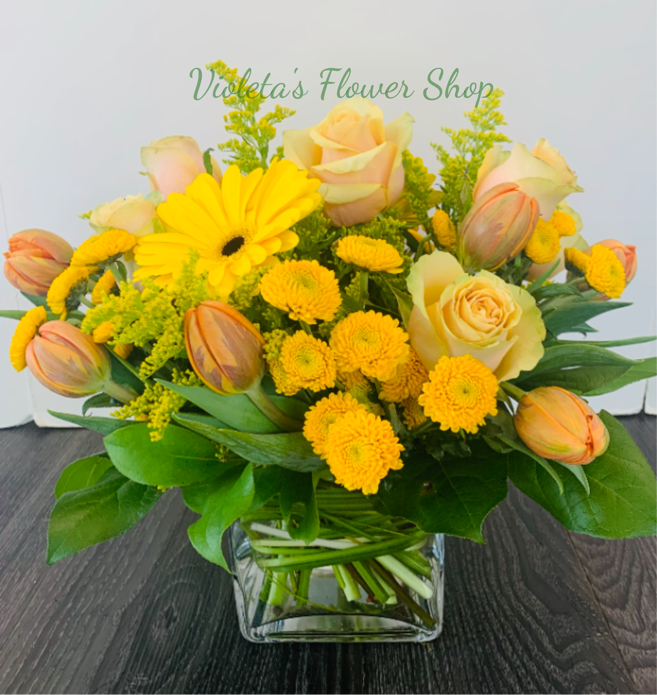 Hello Sunshine Arrangement - Sunlit yellow roses and bright yellow gerberas headline this Hello Sunshine arrangement, perfect as a cheerful birthday bouquet, a spring celebration centerpiece, or a warm thank-you gift. The mix of yellow chrysanthemums and soft peach-and-cream tulips balances sunny energy with a gentle, romantic touch so the person you're thinking of will feel uplifted and genuinely noticed.  Arranged in a clear square glass vase, the low, rounded composition reads fresh and garden-style-loose greenery and broad leaves fill the design for a full, natural silhouette. Expect pale peach roses, yellow button chrysanthemums, cheerful gerbera accents, cream-and-peach tulips and complementary filler blooms in white and pink; the color palette highlights warm yellows with soft blush tones for contrast.  This is the kind of everyday arrangement that brightens a bedside table, office desk, or kitchen counter. Vase-ready and hand-tied for lasting balance, it's sized for easy placement and simple care-keep stems trimmed and fresh water in the clear glass vase to enjoy the blooms longer.