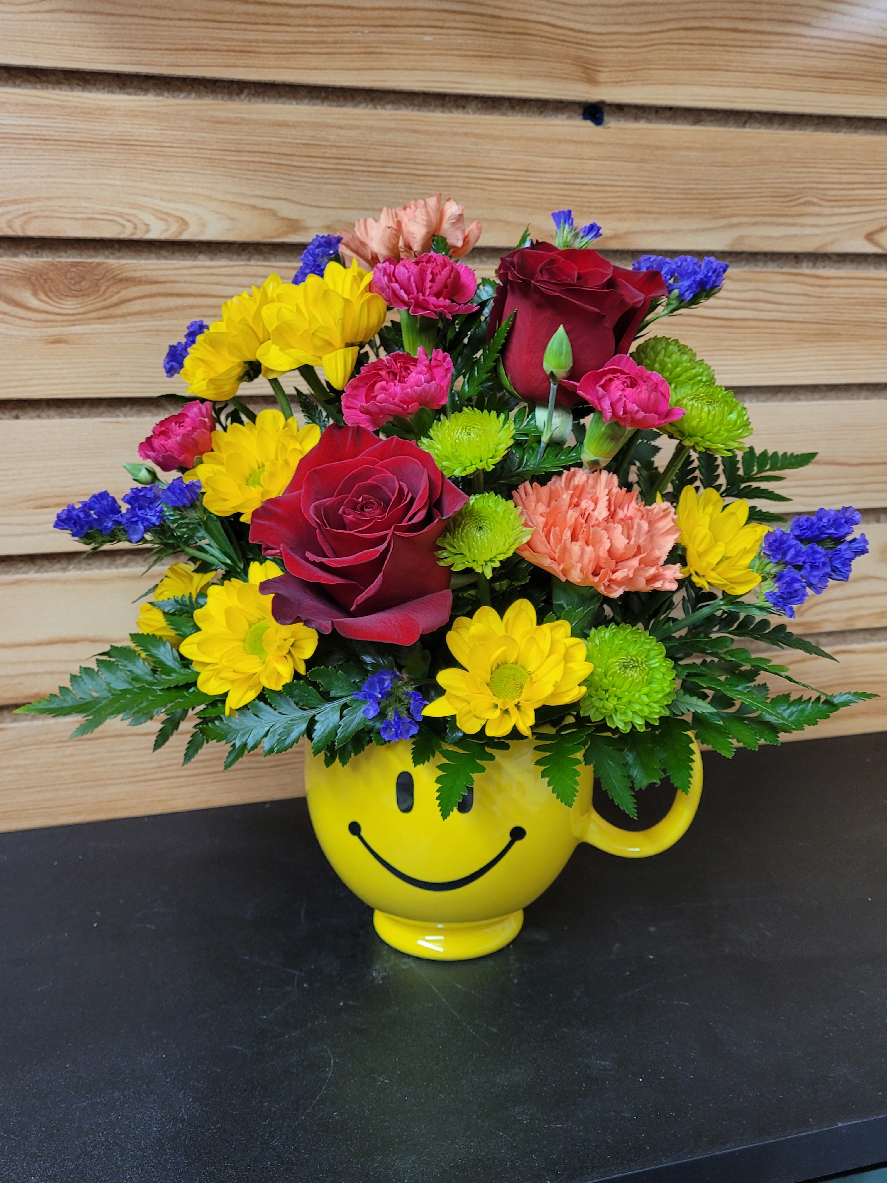 Bright Smiles - A ceramic smiley mug bursting with roses, chrysanthemums, accent flowers, and lush greens. This will bring smiles no matter what the occasion. Flowers may vary based on availability.   