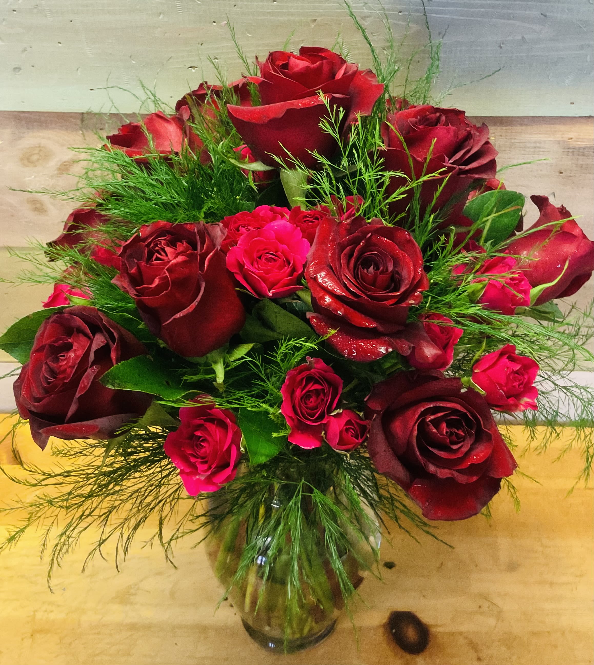 Velvet Petals  - A lush cluster of roses in rich reds and pink-reds creates an immediate impression of romance-perfect as a birthday bouquet, anniversary roses, or a heartfelt Valentine's arrangement. The dense, garden-style grouping reads sumptuous and intimate, the kind of floral gift that makes the person you're thinking of feel deeply cherished.  Deep burgundy to bright pink-red roses are closely gathered with fine, airy green foliage threaded between the blooms for texture and movement. The stems are arranged in a clear glass vase for a classic, elegant presentation that shows off the full heads and leafy stems. The overall look is full and slightly lush, with a warm red palette that reads both timeless and celebratory.  For the recipient, this arrangement sends a clear message of love and admiration-ideal for anniversaries, romantic surprises, or to celebrate someone special. Vase-ready on delivery, it's easy to care for with fresh water changes every few days to keep the roses looking their best.
