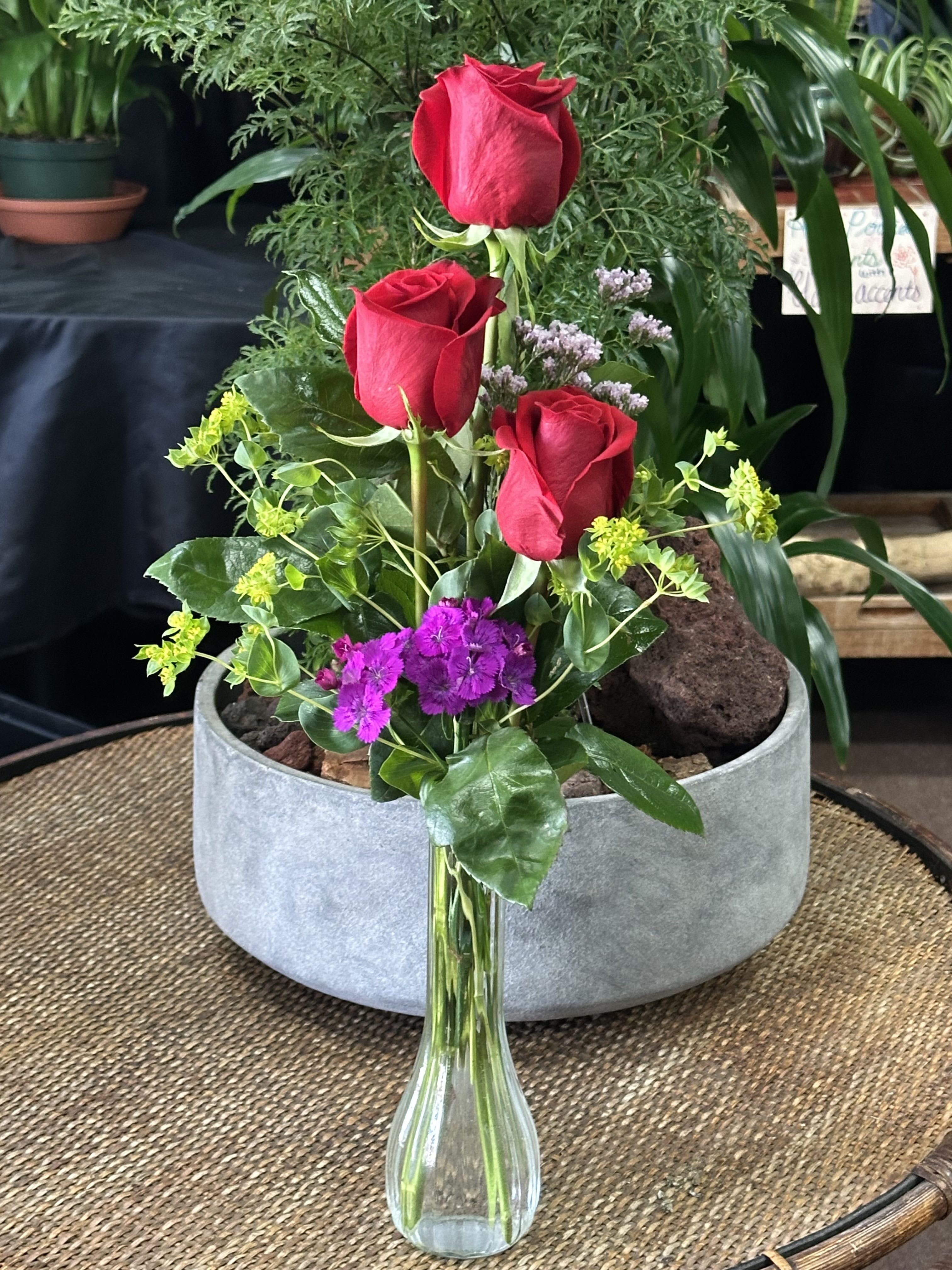 Three red roses in a clear glass vase with purple and yellow accents