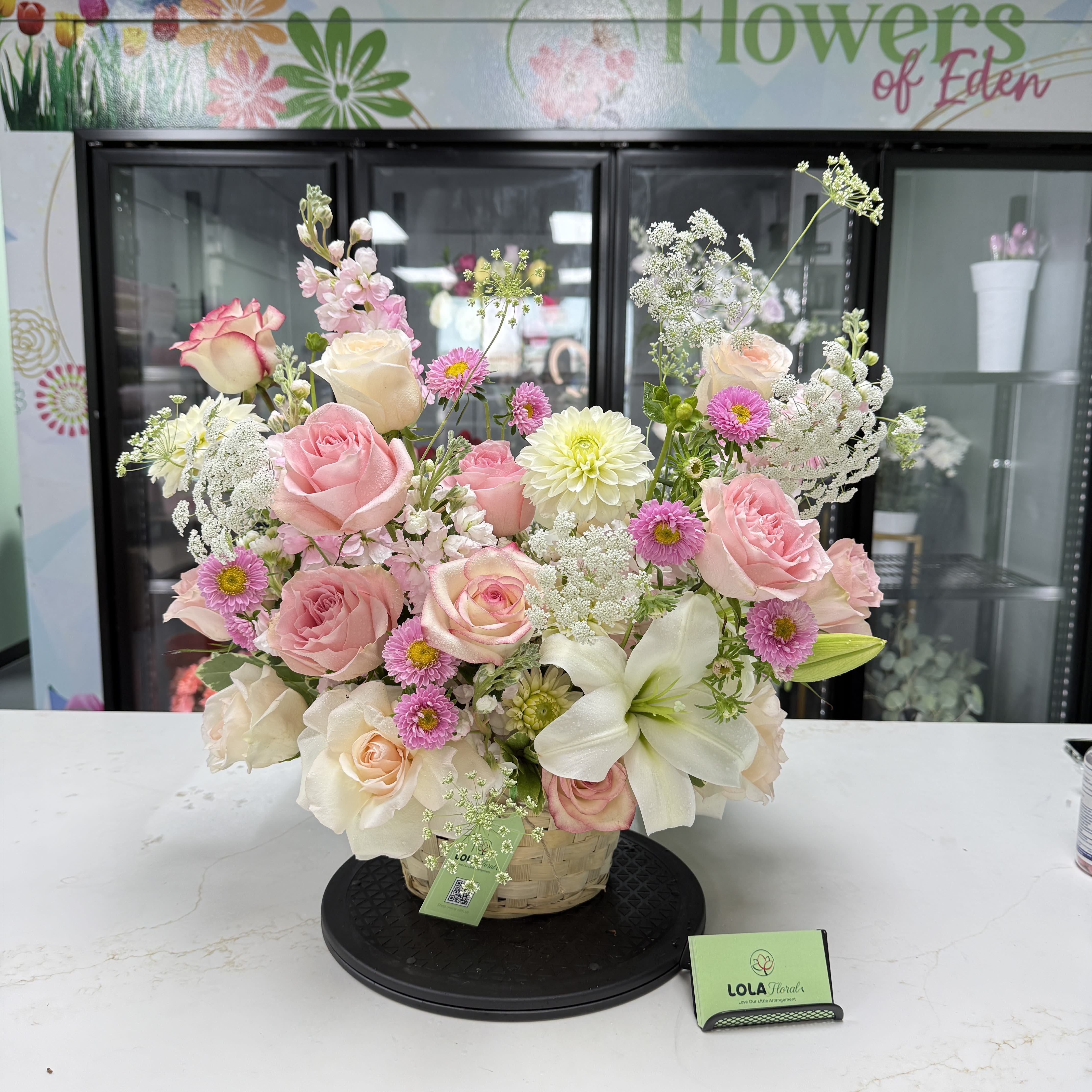 Grand Basket 02 - A soft, garden-style display of pink and white blooms makes this flower arrangement an effortless way to celebrate birthdays, anniversaries, or to say thank you. This flower arrangement offers a fresh, romantic look that will make the person you're thinking of feel cherished-large pink roses and a single white lily create a graceful focal point while the mixed-color palette keeps the mood joyful and bright.  The rounded composition sits in a woven basket and is filled with abundant roses, lilies, daisy-like blooms and airy white filler for texture and gentle height. The loose, abundant styling reads casual yet refined, with green accents that give a natural, just-picked-from-the-garden feel. It arrives on a black display tray and includes a plant tag and business card for easy gifting.  Perfect for spring celebrations or any moment you want to make feel special, this arrangement is the kind of gift that looks as good on a kitchen table as it does on a mantel. Order with a short message and we'll prepare it ready to present-no fuss, just a thoughtful, beautifully arranged bouquet that will brighten their day.