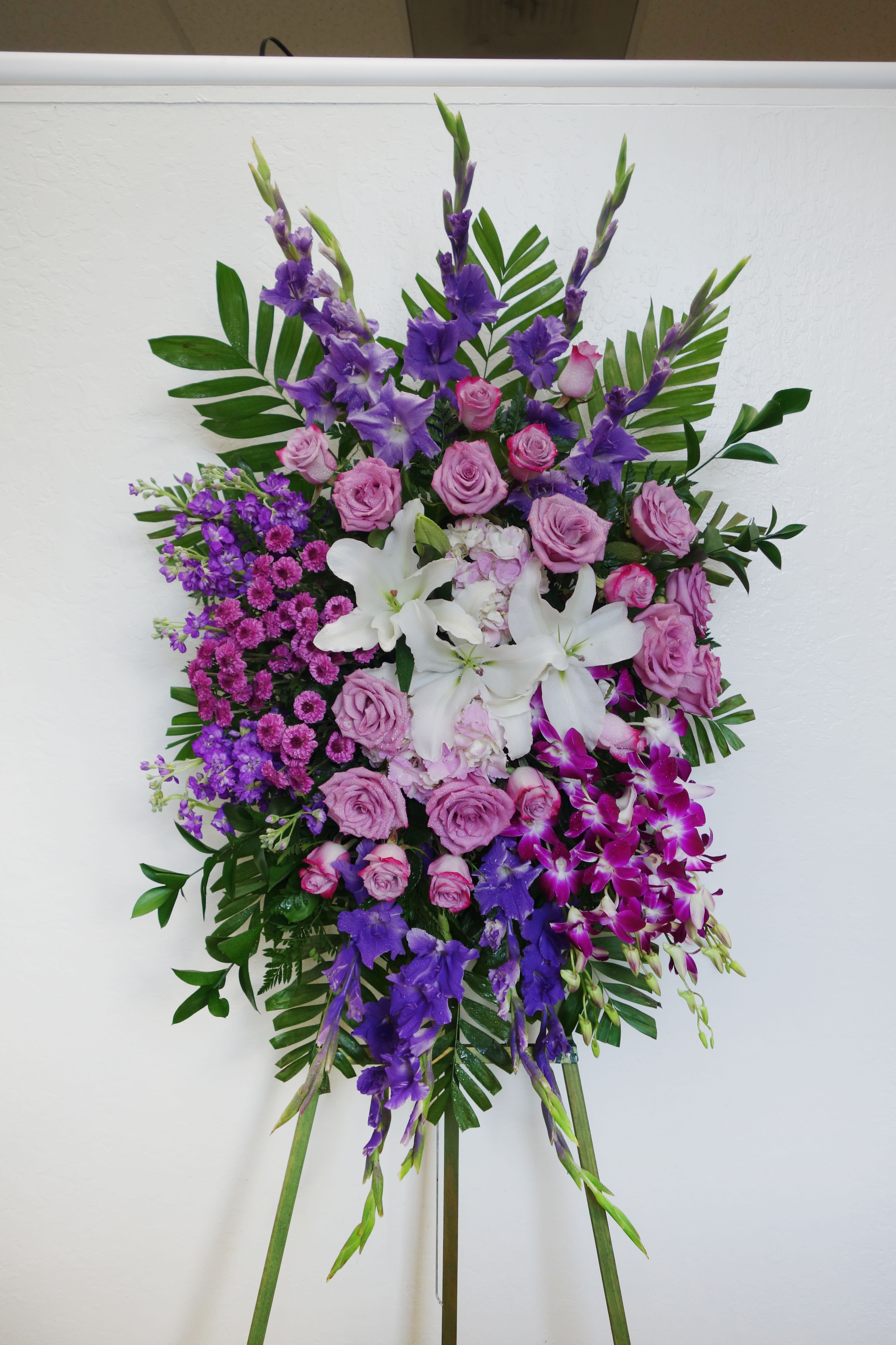 SS#07 Purple Sacred Garden Spray - A lovely lavender spray of flowers lets you share your compassion, hope and beauty with all. Beautifully simple. Beautifully serene. It's the perfect way to send your sincere sympathy. This regal spray includes purple roses, gladiolus, stocks, dendrobium orchids, white oriental lilies, and mixed foliage.  The arrangement is approximately 48" H and 24" W.