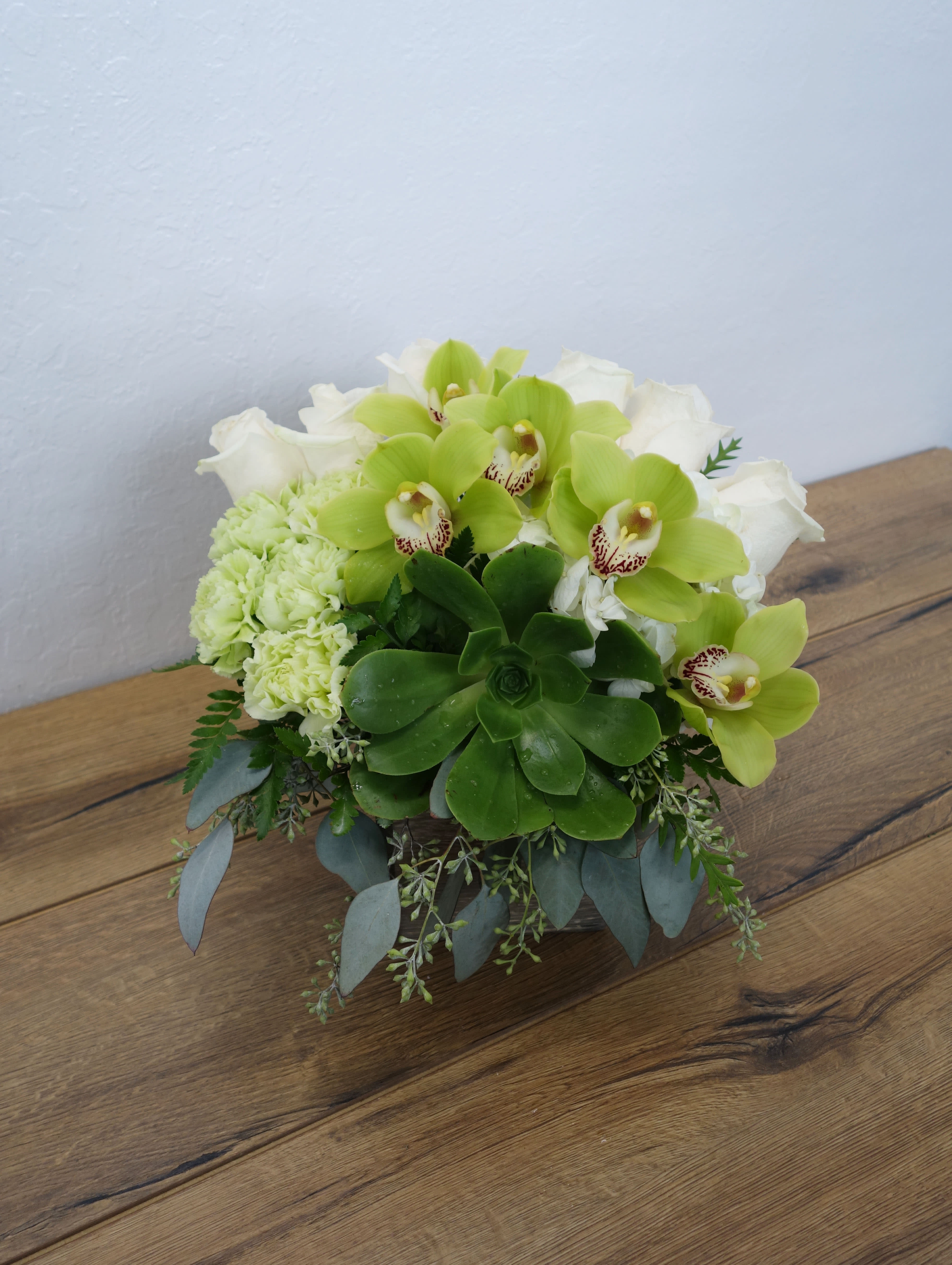 Rainforest Garden - The green abundance of the rainforest is captured in this lovely bouquet brimming with exotic green cymbidium orchids, roses and colorful succulents. Artfully arranged in a glass cube vase, it's a gift that's as right as rain.  A refreshing mix of flowers including green cymbidium orchids, echeveria succulents, roses and carnations accented with eucalyptus in a wooden container.  Deluxe (as shown)