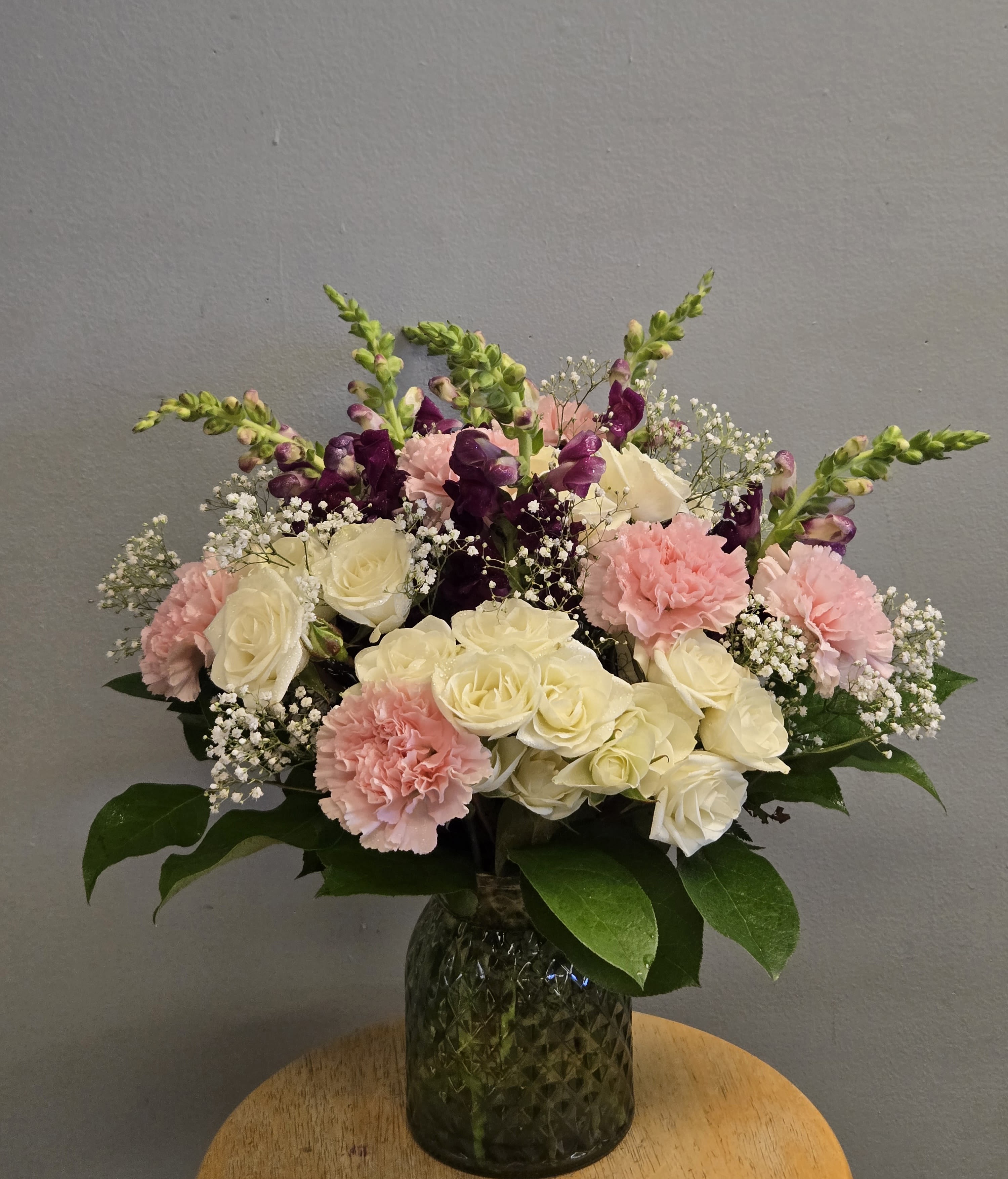 Purple Elegance - This arrangement includes cream roses, pink carnations, purple snapdragons, and delicate white filler flowers arranged with broad glossy green foliage in a dark green glass vase. Perfect for Birthday and Anniversary.