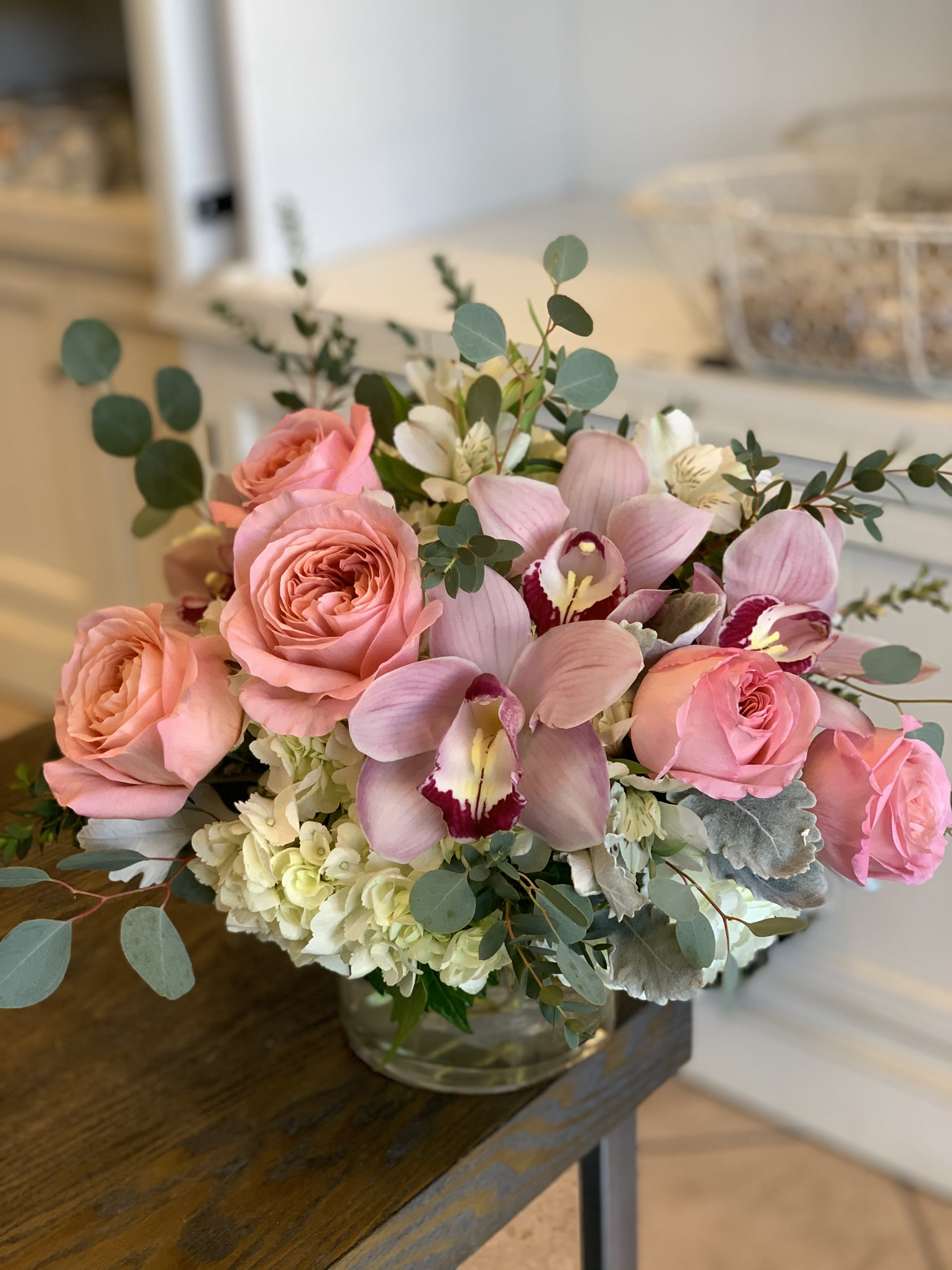 Rustic Charm  - A stunning array of lush hydrangeas, premium pink Expression garden roses, and elegant cymbidium orchids, thoughtfully accented with silver dollar eucalyptus and soft dusty miller for added texture and depth. The garden roses are selected for their naturally full, open bloom and exceptional longevity, offering a romantic, luxurious look from the moment they arrive. This beautifully designed arrangement is perfect for celebrating and honoring someone truly special.