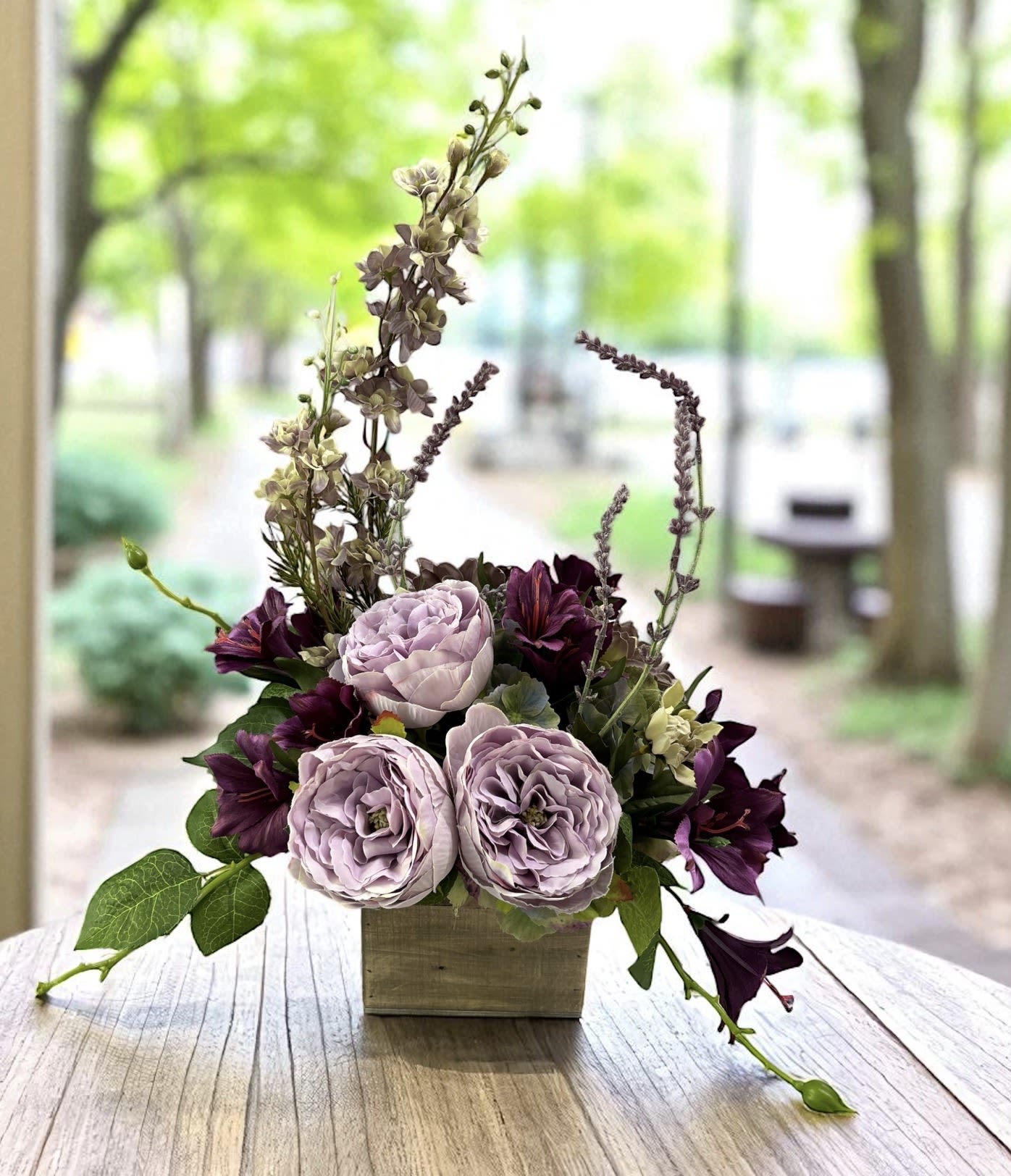 Lavender Serenade Silk Design - Lavender Serenade features lavender garden roses, rich plum blooms, delicate floral accents, and flowing greenery arranged in a charming rustic wooden box. Inspired by the beauty of a flourishing garden, this elegant silk design combines soft lavender tones with deeper plum hues to create a striking display filled with color, texture, and natural movement.  Designed to make a statement without overwhelming a room, Lavender Serenade is perfect for dining tables, coffee tables, entryways, mantels, sideboards, offices, sunrooms, bedrooms, and reception areas. The romantic color palette complements farmhouse, cottage, French country, traditional, rustic, and transitional decorating styles, making it a versatile addition to both home and office décor.  Realistic flowers and lush greenery create the appearance of a freshly gathered garden arrangement while offering the convenience of lasting beauty without watering, pruning, or seasonal replacement. This silk floral design provides year-round color and elegance, making it an easy way to brighten any space.  Lavender Serenade also makes a thoughtful gift for birthdays, anniversaries, housewarmings, retirement celebrations, Mother’s Day, hostess gifts, or anyone who appreciates beautiful floral décor. Flowers, greenery, containers, and accents may vary slightly based on availability while maintaining the overall style, color palette, and character of the design.