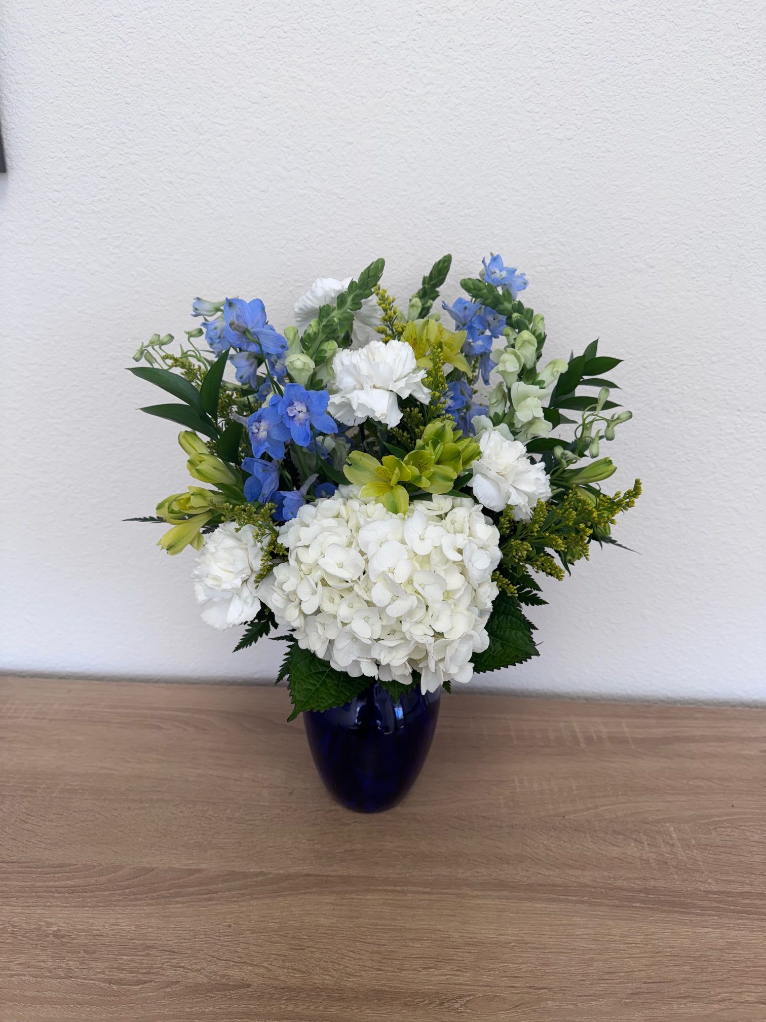Blue Serenity - A cool, calming bouquet anchored by a dense collar of white hydrangeas, Blue Serenity is a flower arrangement perfect for any occasion. The vertical sweep of blue delphinium, white snapdragons carnations, alstroemeria and solidago lift the composition, giving the recipient a sense of freshness and quiet elegance-an arrangement that reads both celebratory and soothing for sympathy moments.  This is the kind of bouquet that will make the person you're sending it to feel seen and comforted-bright enough to uplift, restrained enough to soothe. Ships ready to display in the included blue vase; refresh with clean water and trim stems as needed to extend vase life.