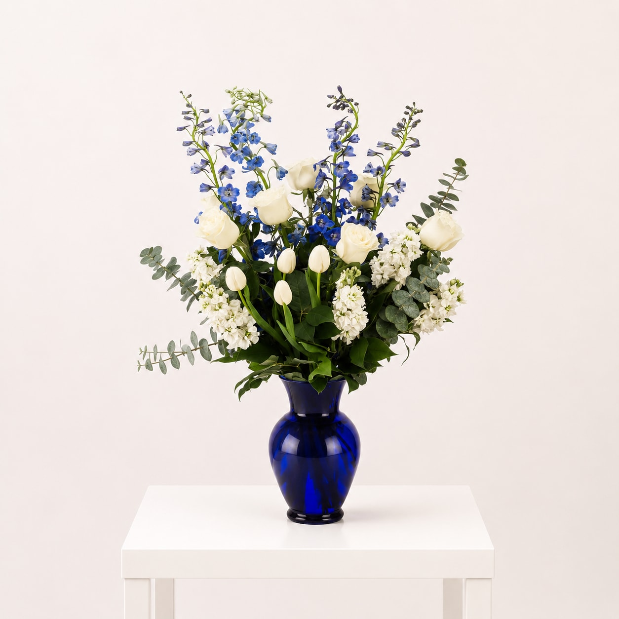 Cobalt Garden  - A striking flower arrangement featuring sapphire-blue blooms, creamy white roses, and fresh seasonal greenery, beautifully designed in a bold cobalt blue vase. Elevated, fresh, and effortlessly refined, this design makes a sophisticated statement in any space.  Each arrangement is Designer’s Choice, thoughtfully created with fresh seasonal blooms. Flower varieties and color tones may vary based on availability while preserving the overall style, palette, and quality.  Perfect for anniversaries, housewarmings, congratulations, Mother’s Day, or sending a meaningful “thinking of you” gesture.  Same day flower delivery available in Southwest Florida, including Naples, Bonita Springs, and Fort Myers Beach.