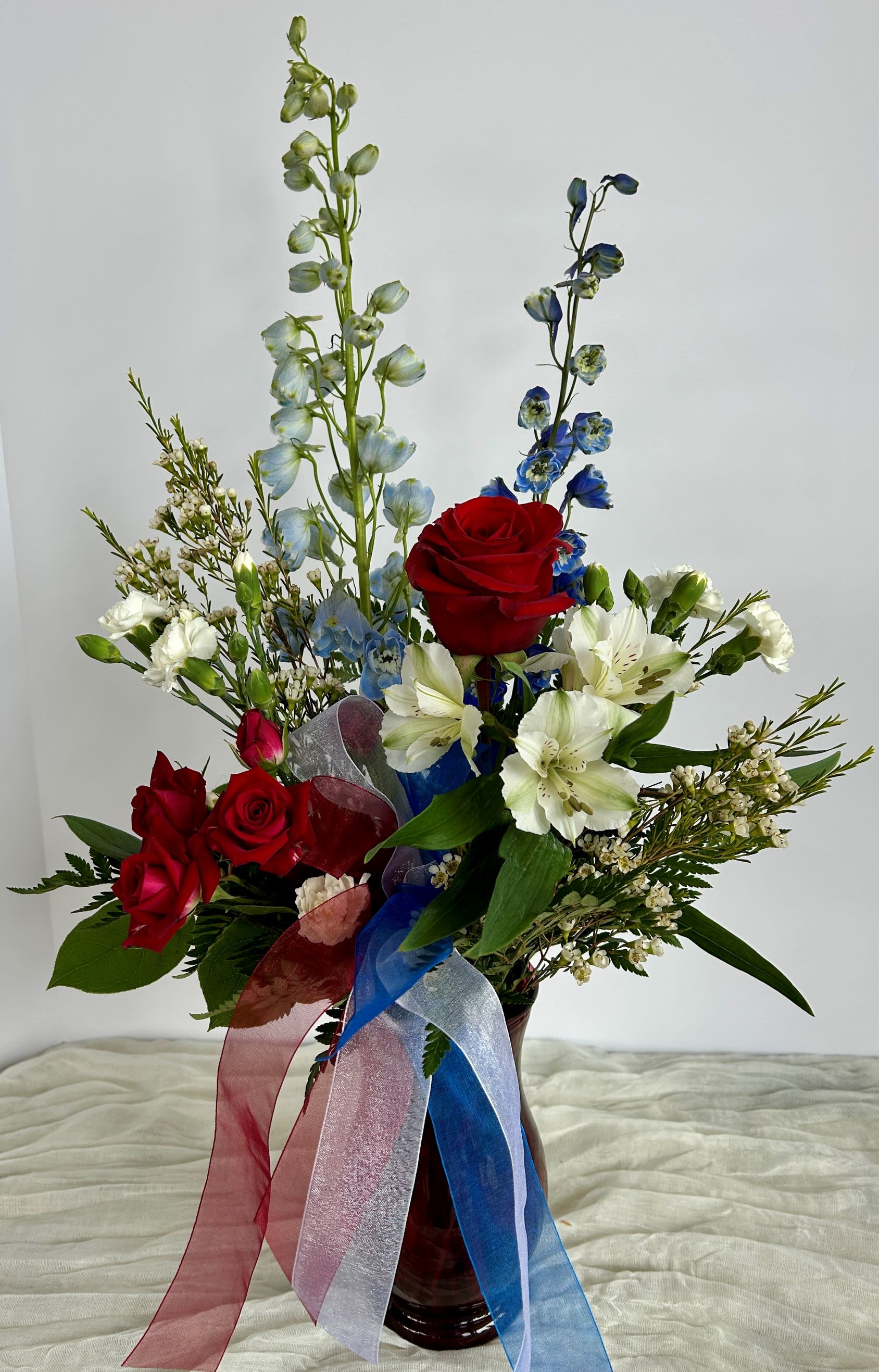 Cheers to the Red, White, and Blue - This patriotic arrangement consists of red roses, white alstroemeria, and blue delphinium. It is filled with wax flowers, mini carnations, and greens, and is styled in a red vase with red, white, and blue ribbons. Share some patriotic cheer, and order your arrangement today.