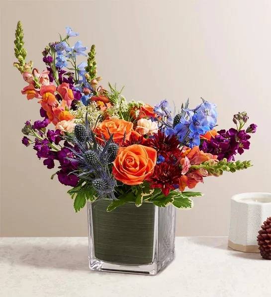 Colorful bouquet of roses, delphinium, and snapdragons in a square glass vase