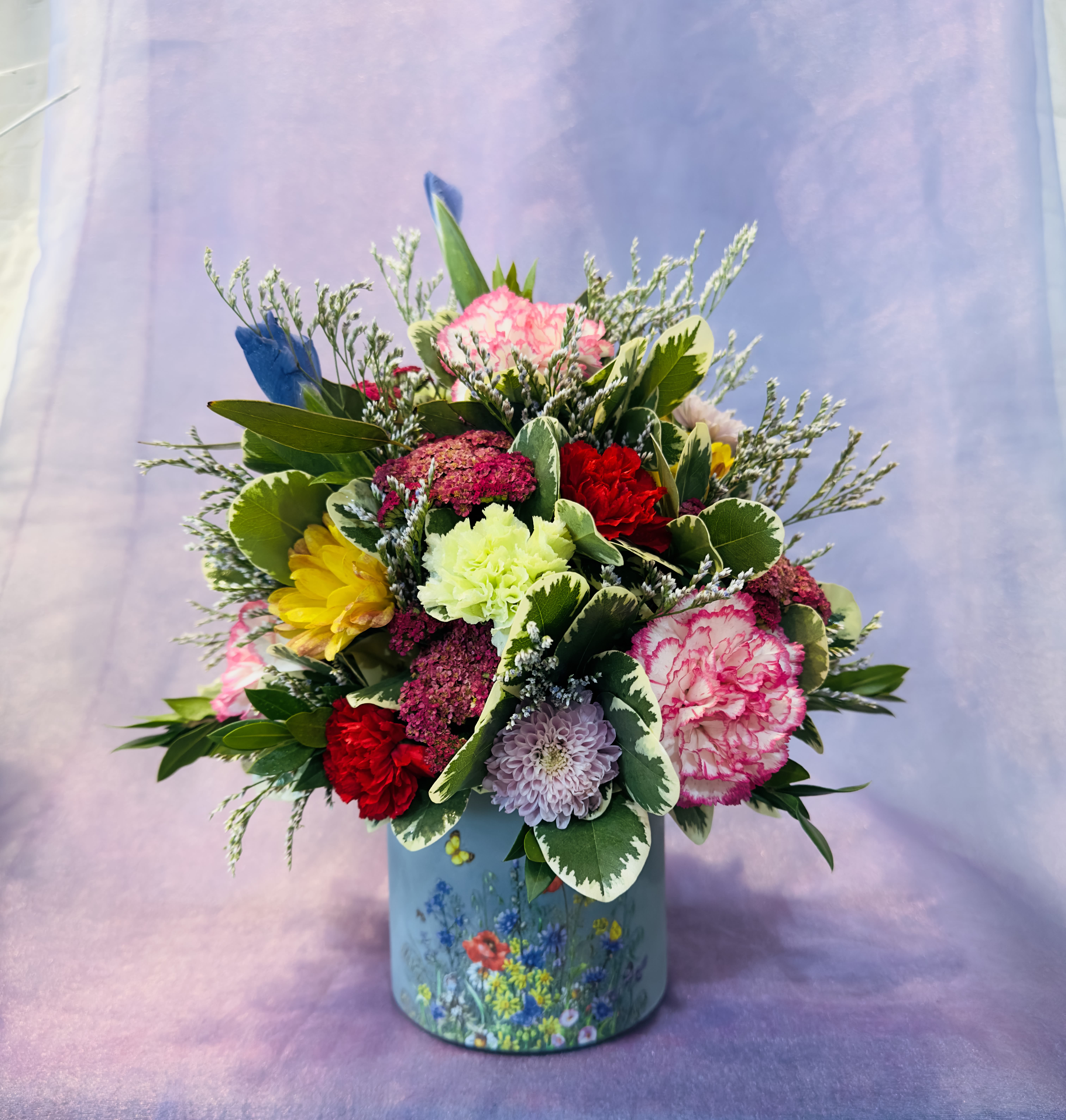 Summer Medley  - A cheerful garden-style arrangement featuring pink and red carnations, pale lavender accents, and sunny yellow flowers nestled among greenery, designed in a decorative wildflower container. The arrangement can change flowers or colors given available flowers during the time period.