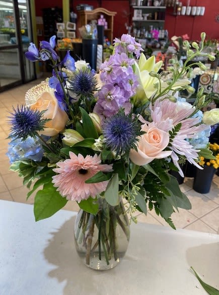 Pastel mixed bouquet with roses, gerbera daisies, and blue accents arranged in a clear glass vase