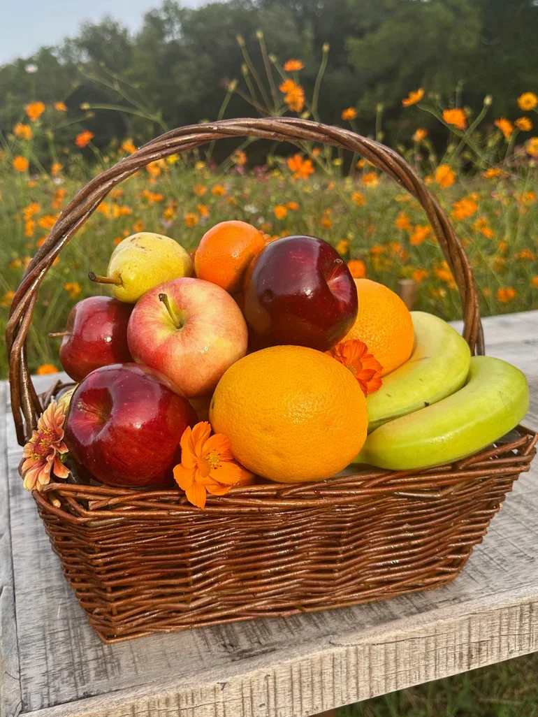 Wicker basket filled with assorted fresh fruits and a few orange blossoms on a rustic wooden surface
