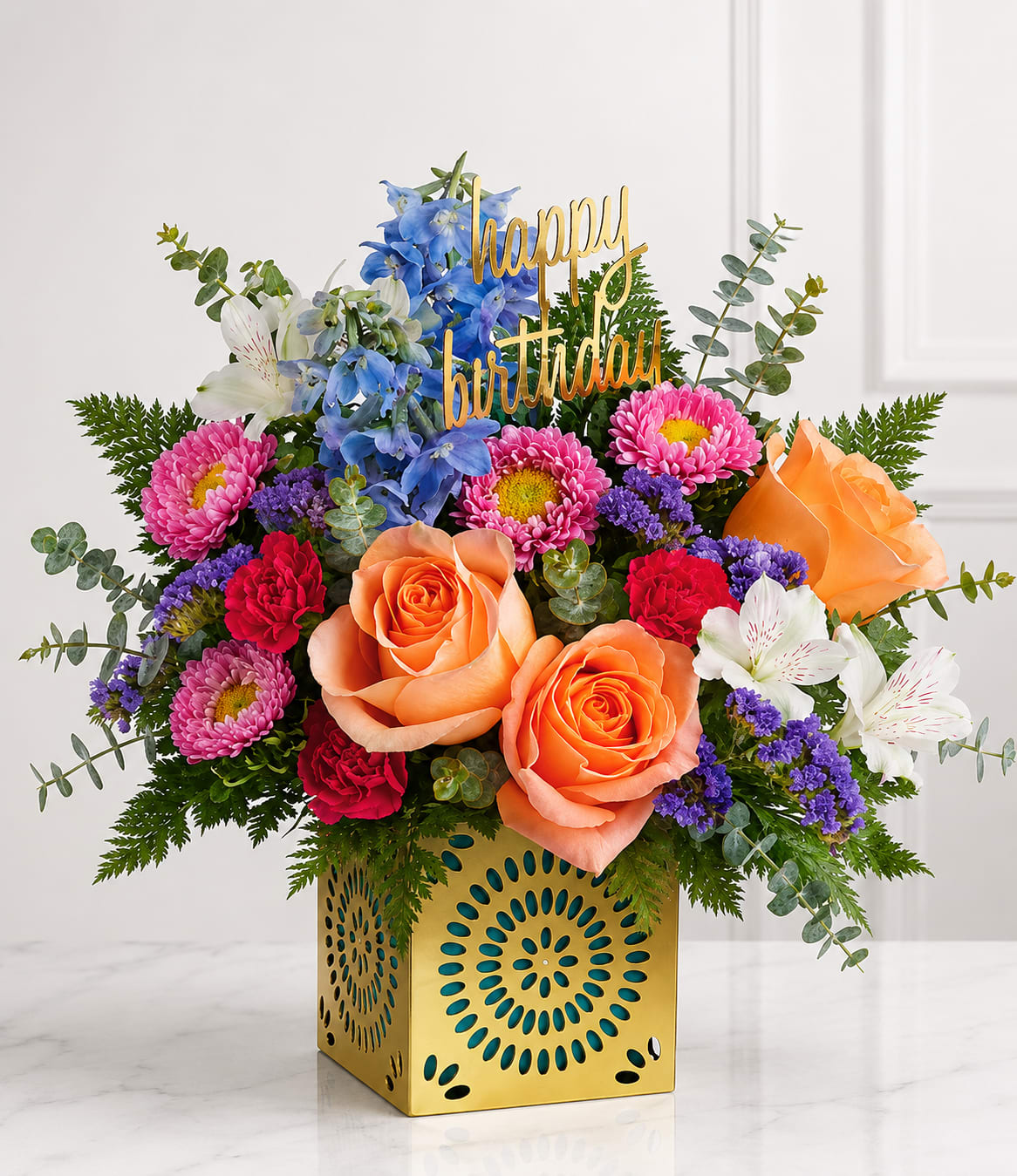 Golden Celebration Bloom Cube™ - Celebrate their special day with the Golden Celebration Bloom Cube™, a vibrant floral arrangement designed to brighten any birthday. Featuring premium peach roses, blue delphinium, pink chrysanthemums, white alstroemeria, red carnations, purple statice, fresh eucalyptus, and lush seasonal greenery in a decorative gold keepsake cube, this arrangement is finished with a cheerful "Happy Birthday" topper. It's a joyful gift that brings elegance, color, and lasting smiles to any celebration.