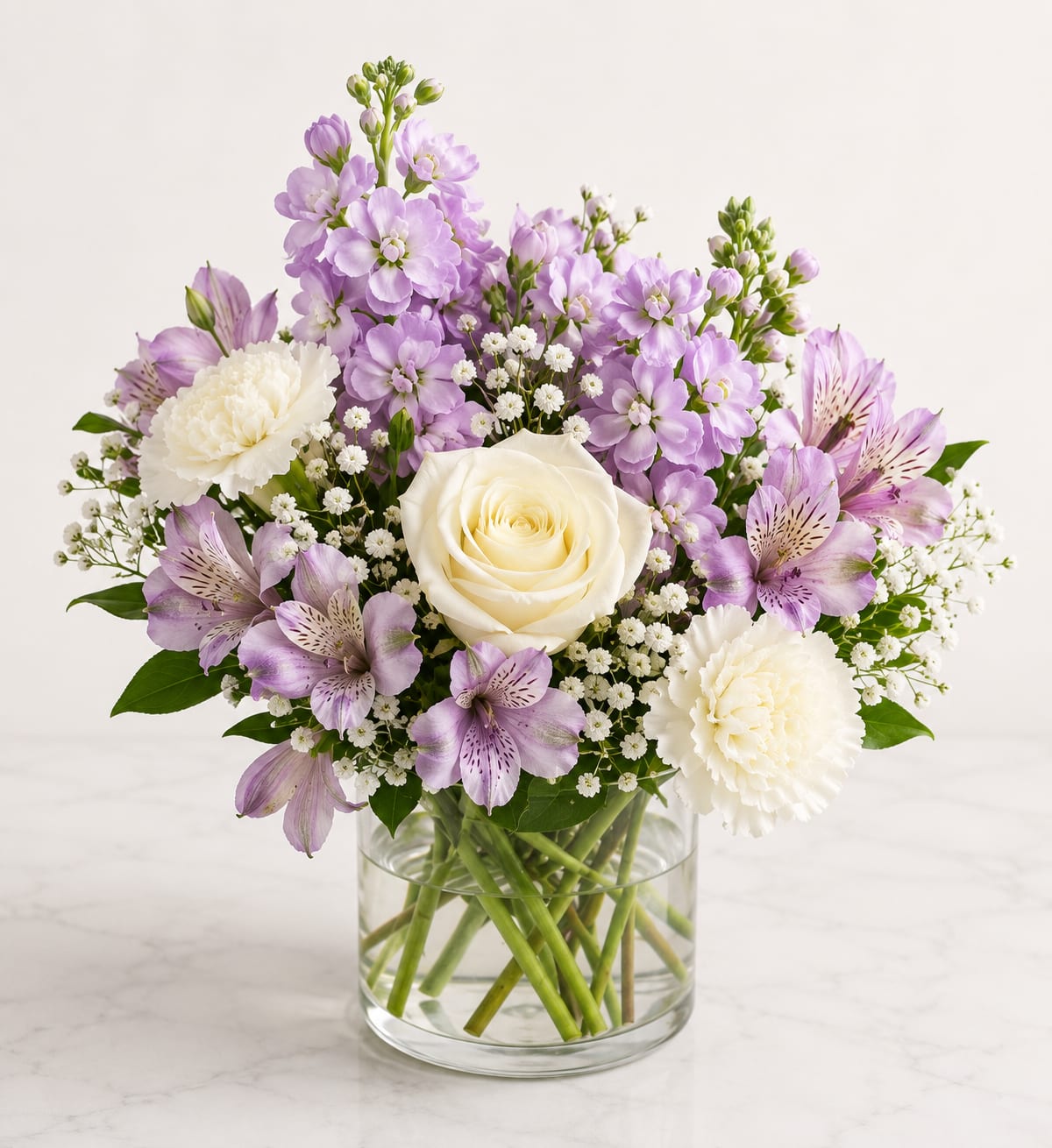 Lavender Grace Bouquet™ - Elegant, peaceful, and timeless, Lavender Grace Bouquet™ showcases a beautiful blend of creamy white roses, delicate lavender stock, graceful alstroemeria, white carnations, airy baby's breath, and fresh seasonal greenery, all artfully arranged in a classic clear glass vase. Its soft pastel palette makes it a thoughtful gift for birthdays, anniversaries, sympathy, get well wishes, thank-you gifts, or simply expressing love and appreciation with grace.