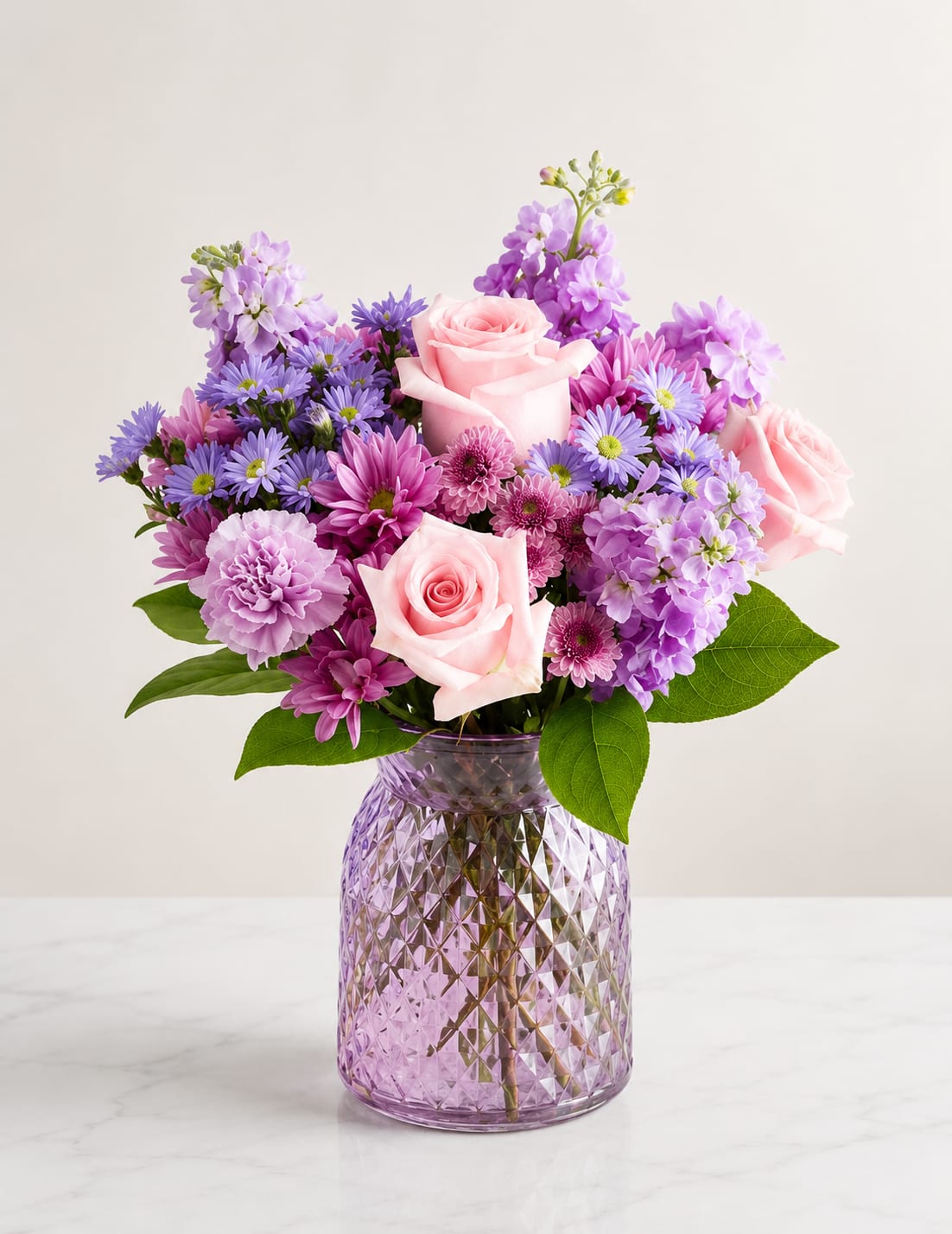 Lavender Blush Garden - A charming garden-style bouquet featuring soft pink roses, lavender stock, purple daisies, carnations, and vibrant chrysanthemums arranged in a beautiful lavender textured glass vase. Designed to bring elegance, warmth, and cheerful color to any celebration or everyday moment.