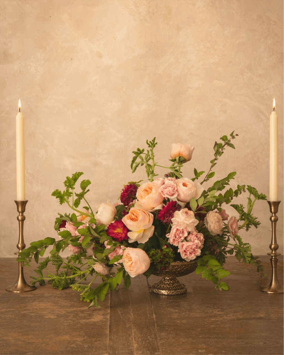 Low floral centerpiece with blush and peach roses on a pedestal, flanked by two lit taper candles