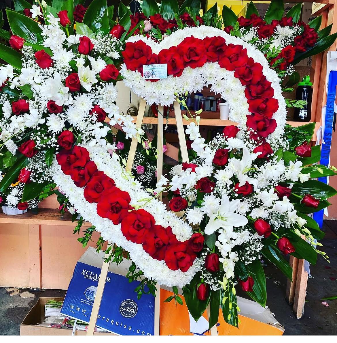 Open red and white heart with burst - A large heart-shaped funeral spray crafted on a sturdy wooden easel, this open red and white heart with burst makes a powerful tribute for a memorial or sympathy service. The vivid red rose border framed by dense white blooms creates an immediate visual contrast-the kind of arrangement that reads as both romantic and reverent, conveying love and respect for the person you're honoring.  Inside the heart, white lilies, chrysanthemums, daisies and sprays of orchids and baby's breath are layered to create a full, formal composition with lush green accents for depth. The structured design and generous scale give the piece strong presence at a viewing or graveside, with textures ranging from the smooth roses and lilies to the delicate airy clusters of baby's breath.  Designed for remembrance, this heart-shaped arrangement communicates devotion and consolation to the person you've lost and to those gathered with them. Delivered ready to display on the included wooden easel; no assembly required.