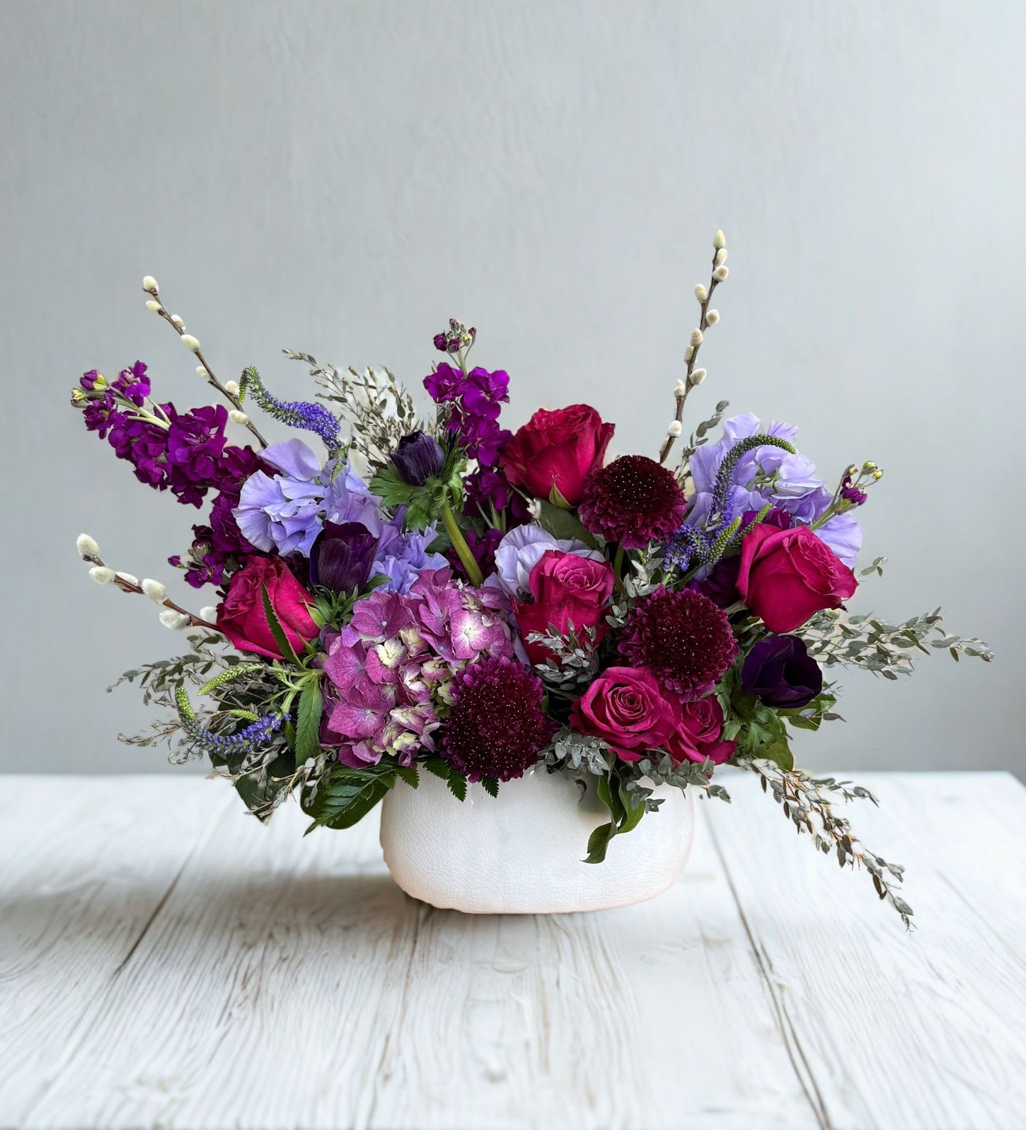 Garden of Gratitude - A lush blend of rich jewel tones and soft lavender hues, this elegant arrangement brings together roses, hydrangea, and seasonal blooms in a low, modern vessel. Accented with delicate textures and airy greenery, it offers a perfect balance of bold color and graceful movement. Designed to make a statement, this piece is ideal for celebrations, heartfelt gifts, or adding a touch of sophistication to any space.