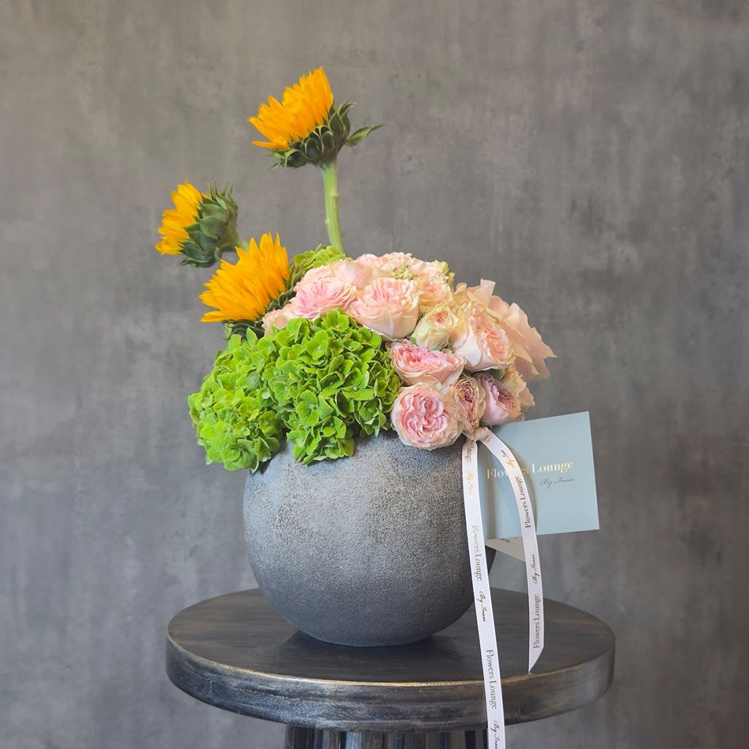 Golden Blush - A radiant arrangement of blush roses and fresh green hydrangea, accented with vibrant sunflower blooms, designed in a modern ceramic vase. Bright yet refined, this piece blends warmth and softness for a fresh, uplifting look—perfect for celebrations or thoughtful gifting. Custom greeting card included.