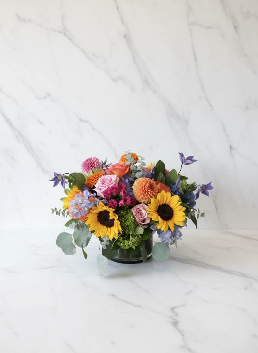 Mixed bouquet with sunflowers, roses, and purple blooms in a low vase