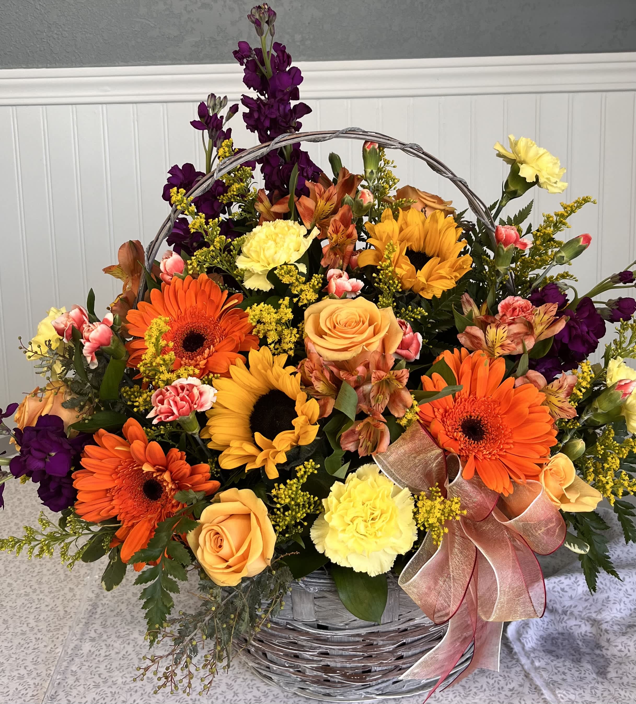 Basket arrangement of orange and yellow flowers with purple accents and a ribbon