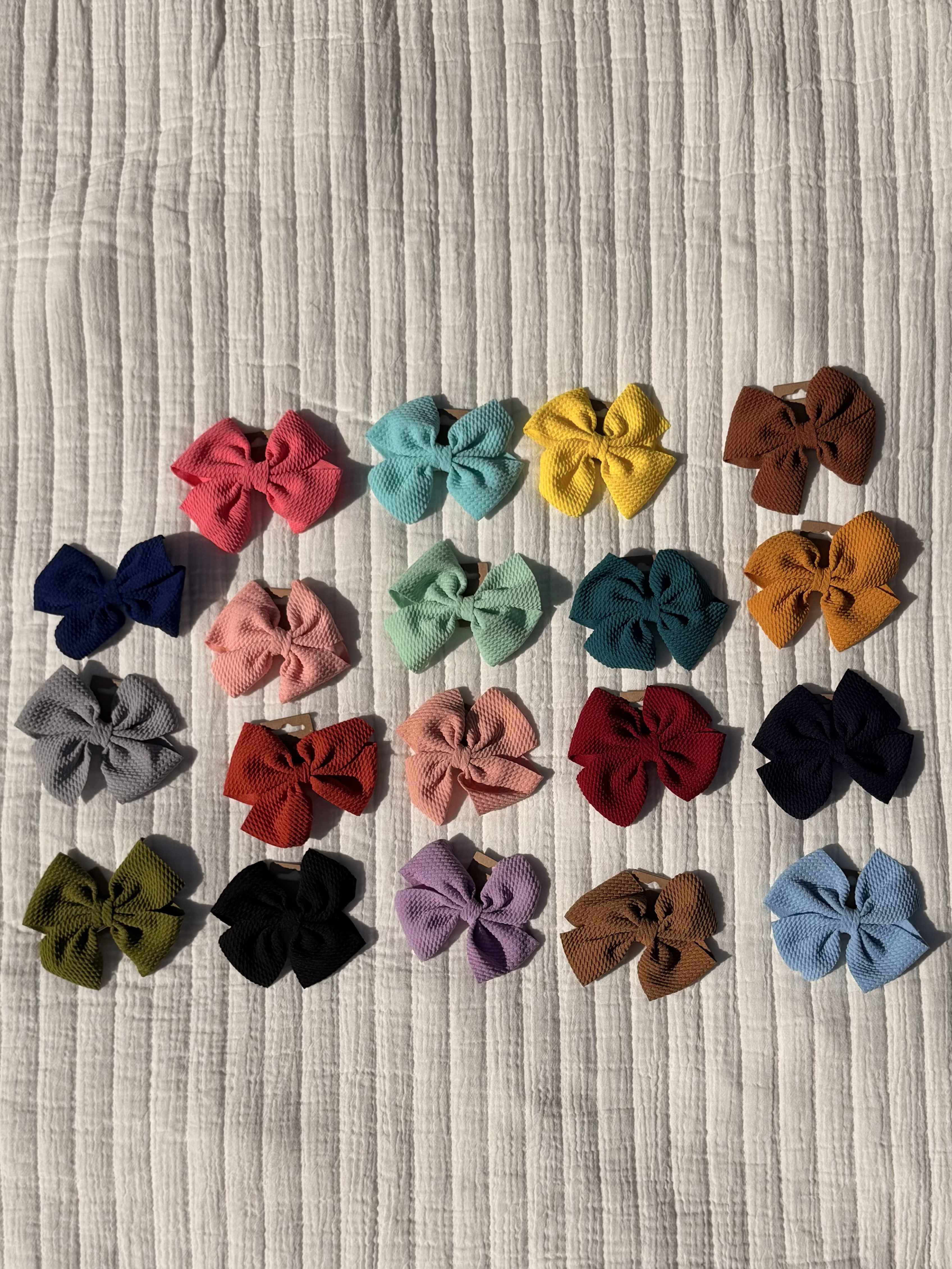 Hand made Hair Bows for baby or for toddlers - Each bow is crafted from slightly structured textured fabric and mounted on a secure hair clip for easy use on fine baby or toddler hair; they are shown laid out in neat rows to highlight the range of solid colors and tidy, handcrafted finish. The compact size and sturdy clip make them practical for active little ones while still feeling delicate and polished.  Great as a single thoughtful surprise, part of a gift set, or a small Mother's Day or Women's Day treat for a mom who loves coordinating accessories, these bows help the person you're thinking of feel noticed and adorable. Order sets to mix-and-match across outfits; care is simple-gently spot-clean when needed and avoid soaking so the clips stay working and the fabric keeps its shape.
