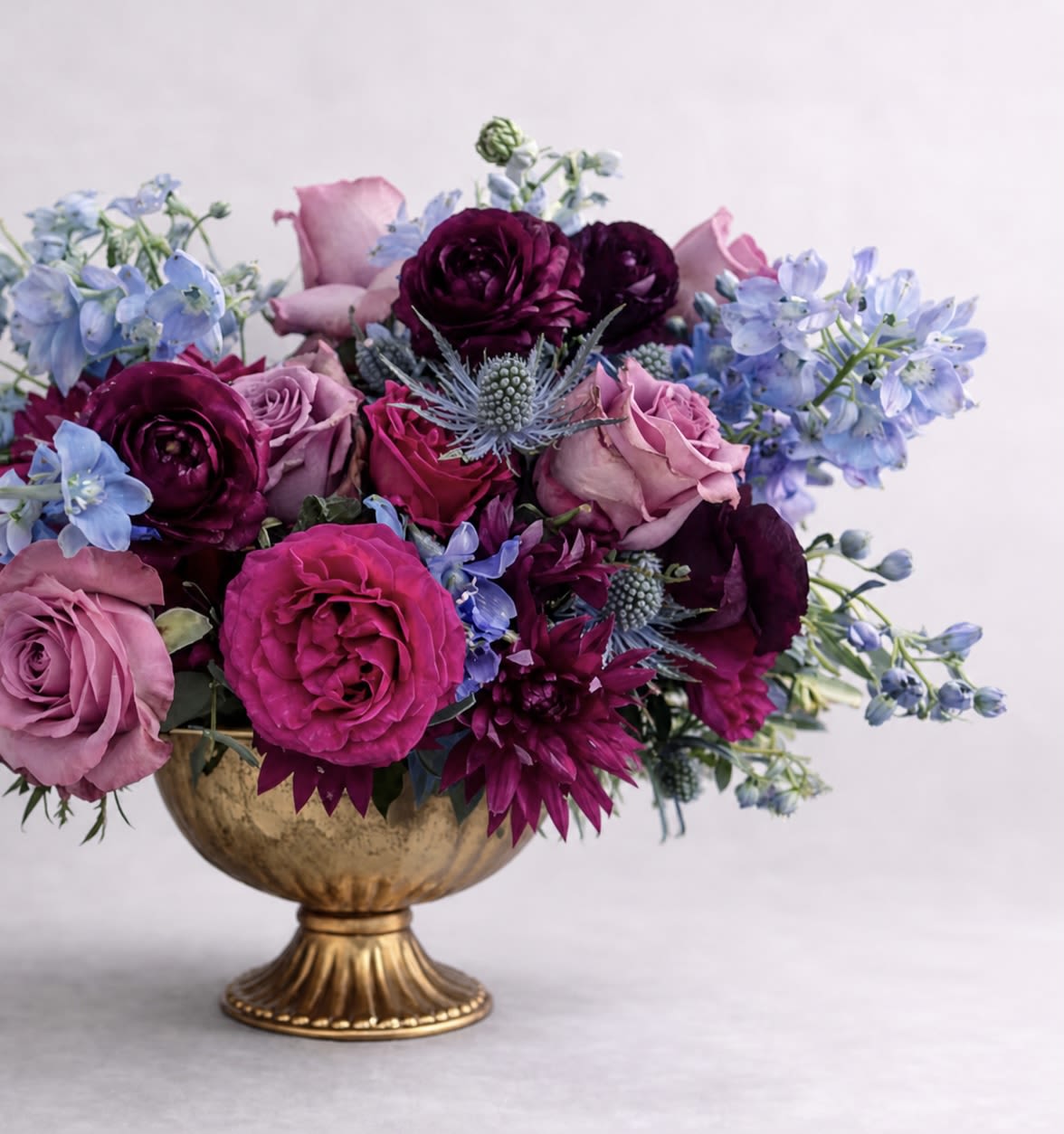 Chef's Kiss - Immerse yourself in the captivating beauty of our April May Flowers original cult favorite and top-selling arrangement, the Chef’s Kiss. This stunning floral masterpiece showcases a harmonious blend of moody gem tones that evoke a sense of mystery and allure. Lush blue delphinium cascades gracefully, their vibrant hue reminiscent of twilight skies. Nestled among them are exquisite purple roses, symbolizing love and enchantment, while rich purple mums add texture and depth. Completing this luxurious arrangement are delicate ranunculus, their soft petals bringing a touch of whimsy.  Perfect for any occasion, whether it’s a romantic gesture or a celebration of friendship, our Chef’s Kiss design is made to impress. Its rich colors and varied textures create a striking visual impact that will leave a lasting impression. Elevate your gifting experience with this enchanting arrangement that captures the essence of elegance and sophistication. 