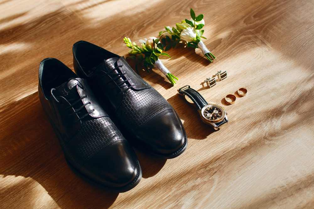 Black dress shoes with boutonniere, cufflinks, rings, and watch on wood floor