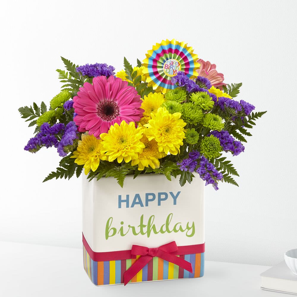 Birthday Brights Bouquet - The Birthday Brights Bouquet is a true celebration of color and life to surprise and delight your special recipient on their big day! Hot pink gerbera daisies and orange roses take center stage surrounded by purple statice, yellow cushion poms, green button poms, and lush greens to create party perfect birthday display. Presented in a modern rectangular ceramic vase with colorful striping at the bottom, "Happy Birthday" lettering at the top, and a bright pink bow at the center, this unforgettable fresh flower arrangement is then accented with a striped happy birthday pick to create a fun and festive gift.