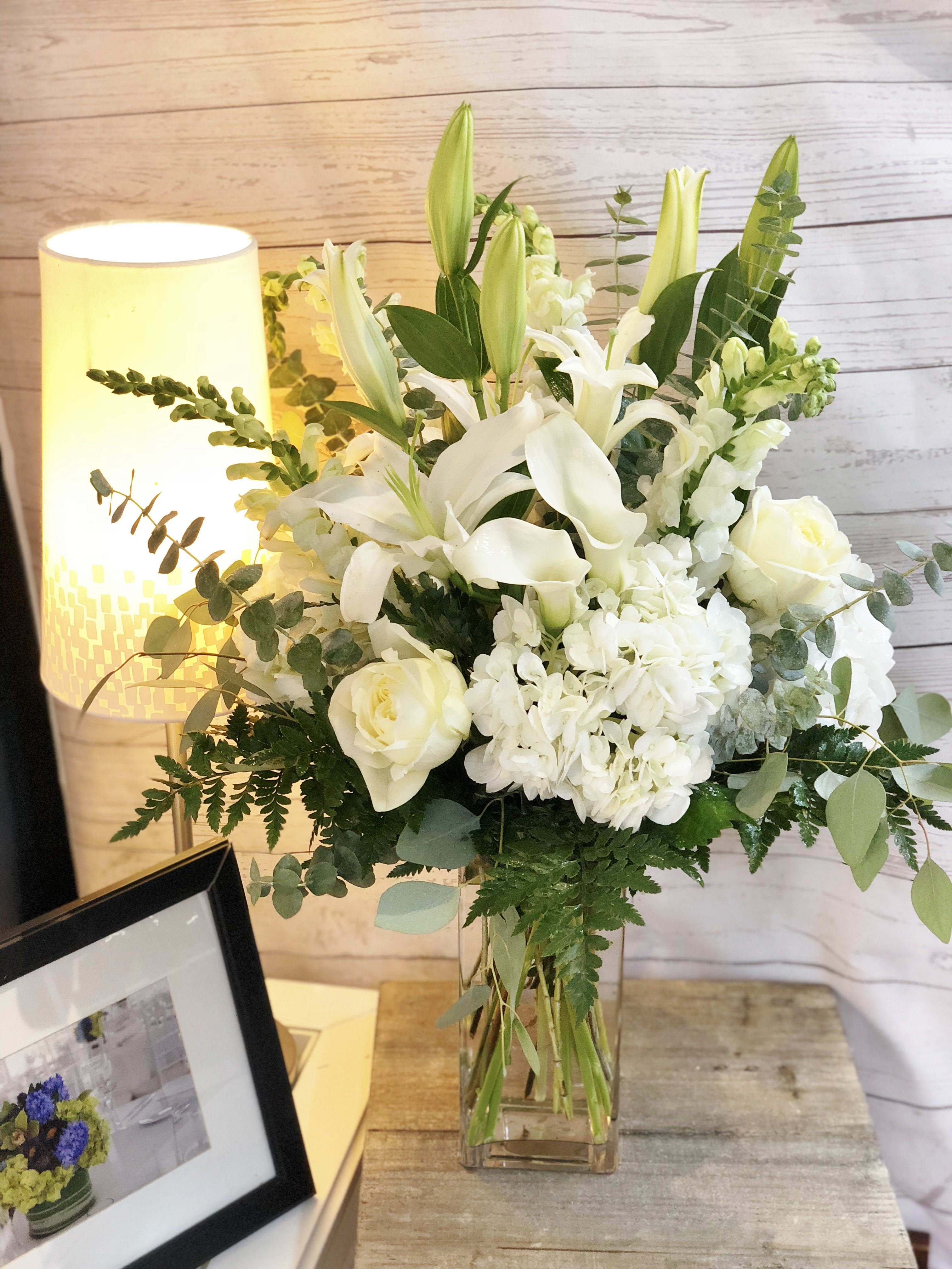 Tall white lily, rose, and hydrangea arrangement with greenery in a clear glass vase on a wooden table