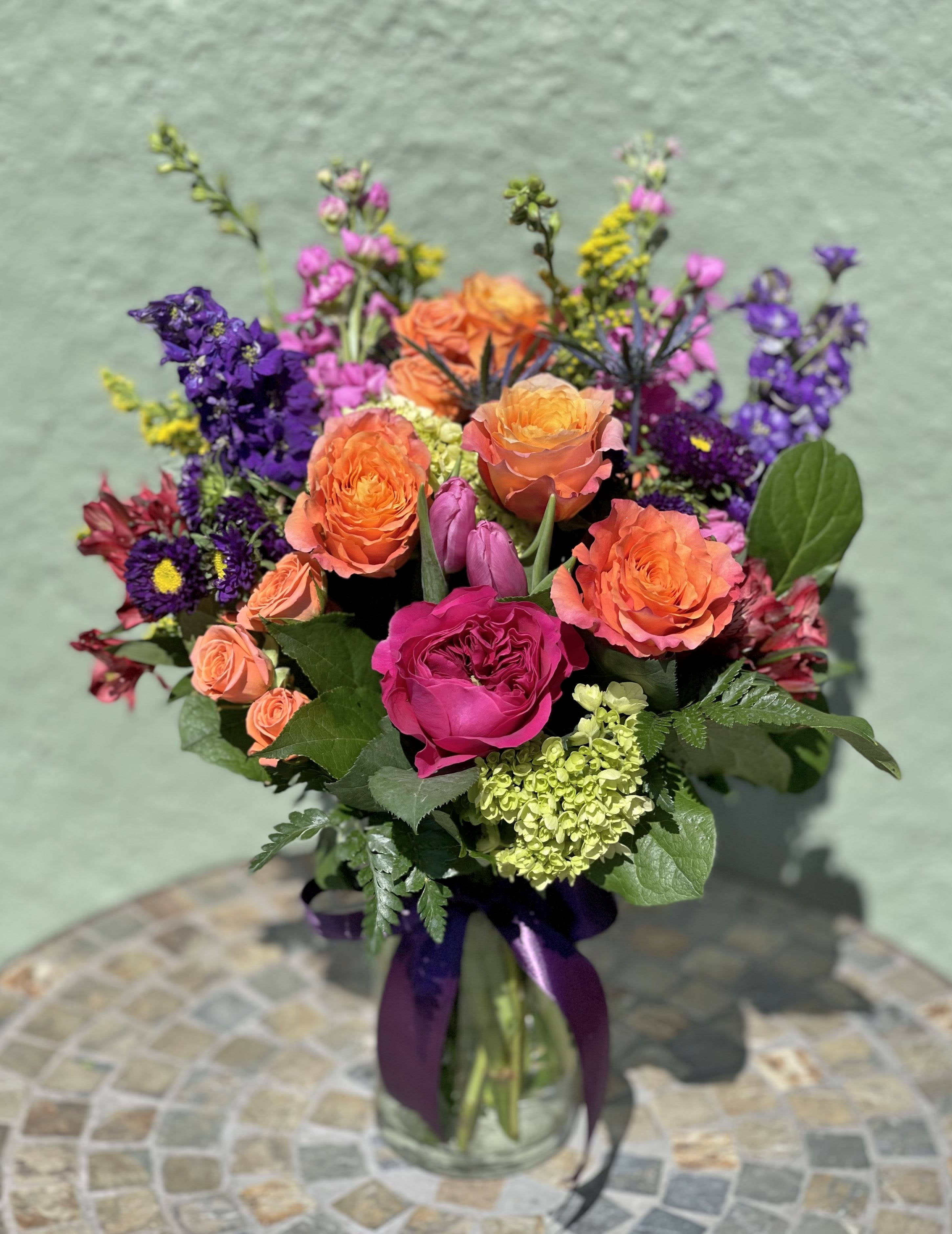 Bright mixed bouquet of orange roses, pink blooms, and purple flowers in a glass vase with a purple ribbon