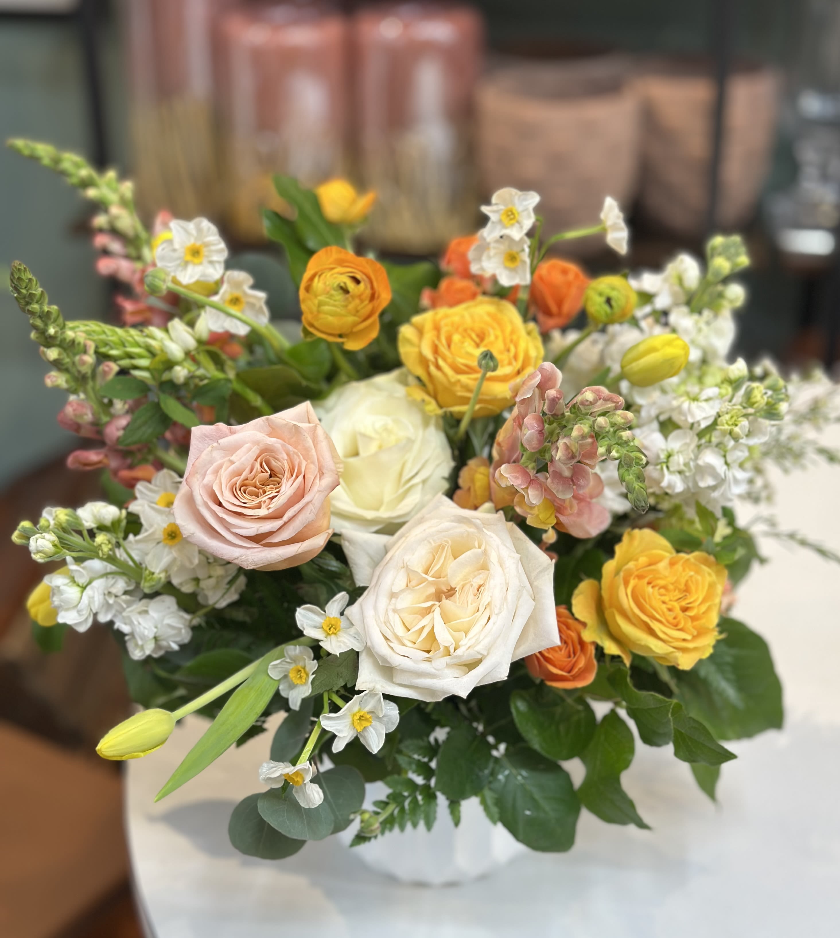 Mixed arrangement of yellow, peach, and white roses with spring blooms in a white vase
